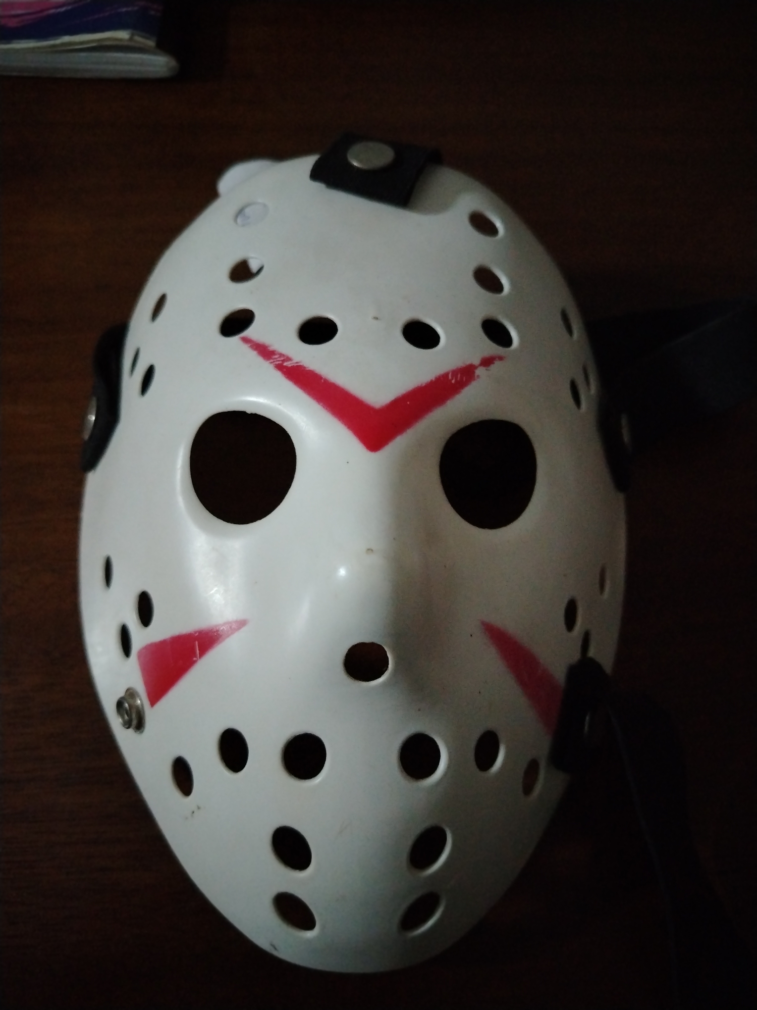 A white, plastic hockey mask with circular vent holes and red chevron markings is centered in the frame. The mask has two large, black eyeholes, a small hole for the nose, and black straps attached to the sides and top. It rests on a dark, possibly wooden, surface. The lighting is dim, creating shadows around the mask. No people are visible. The city of Tinaquillo, Venezuela is not verifiable from the image.