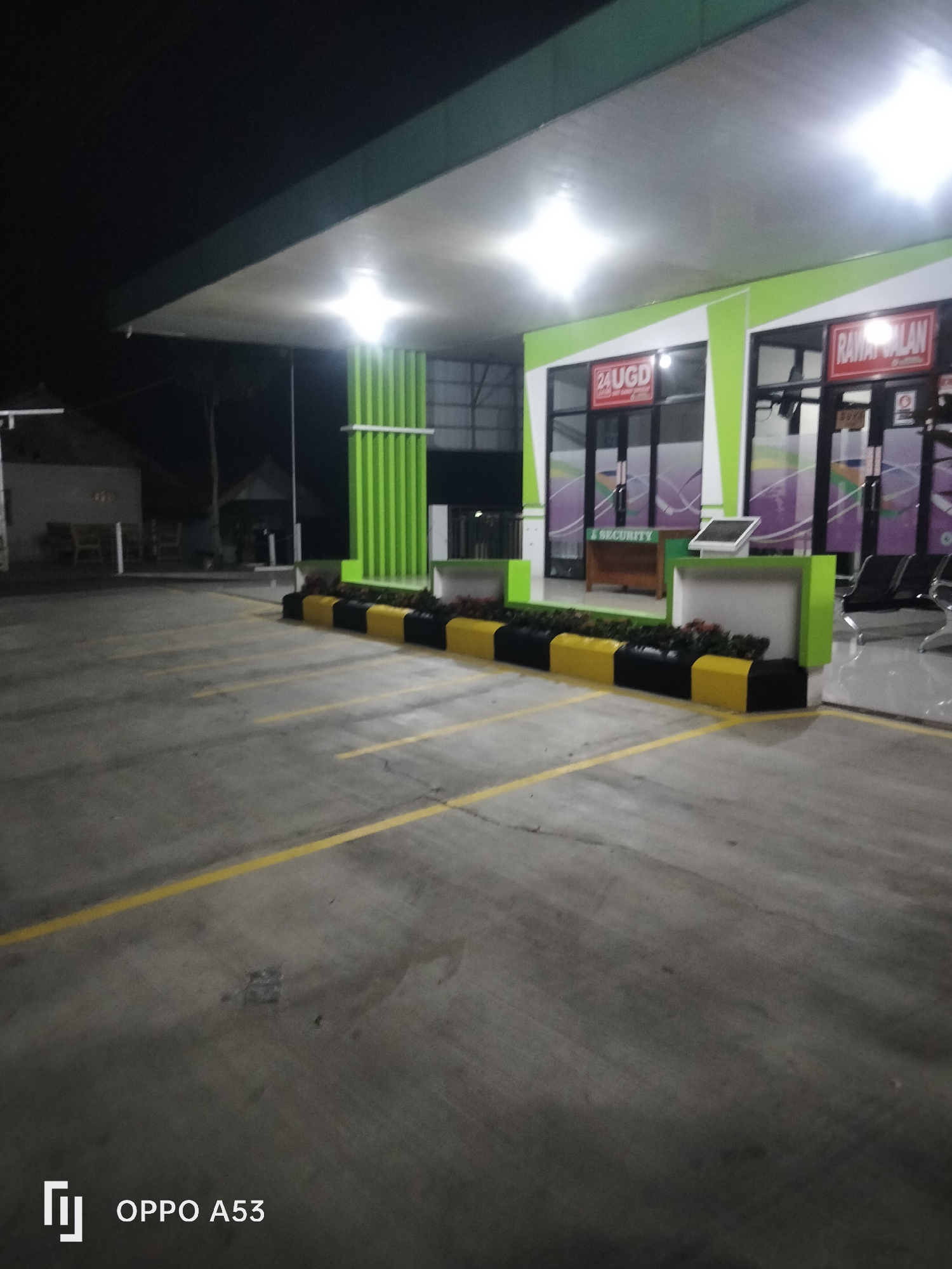 The image shows the exterior of a building at night, identified as the Emergency Room (UGD) and Inpatient Care (Rawat Inap) of a facility in Karawang, Indonesia. The building features a green and white facade with signage indicating "24 UGD" and "RAWAT INAP." The entrance appears to be a glass door. In front of the building, there is a parking lot with yellow lane markings. A low barrier, painted with alternating black and yellow sections, separates the parking lot from a landscaped area with plants. The scene is illuminated by artificial lighting, casting a bright glow from the building's interior and overhead lights. A security desk is visible inside, and there are benches for waiting outside. The overall environment suggests a medical facility operating during nighttime hours.