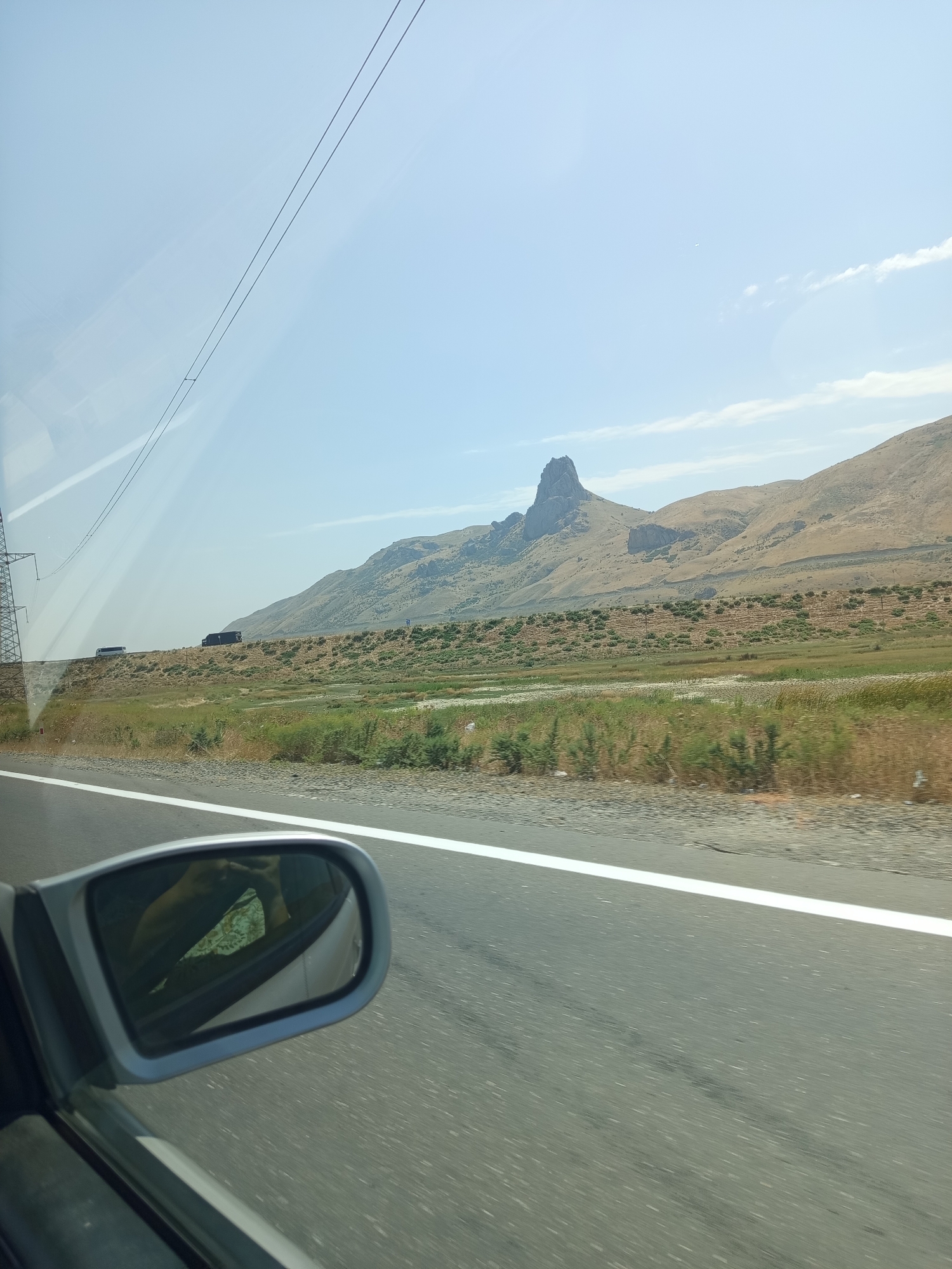 A side view from a vehicle window reveals a mountainous landscape under a clear sky. A distinctive, jagged rock formation rises prominently in the distance, set against rolling hills of dry, sparse vegetation. A highway with a white line marking the lane runs across the foreground, with the side-view mirror of the vehicle visible in the lower left corner. In the far distance, power lines and a transmission tower can be seen, along with a few distant vehicles on what appears to be a road. The scene captures a sense of travel through a rugged, arid environment.
