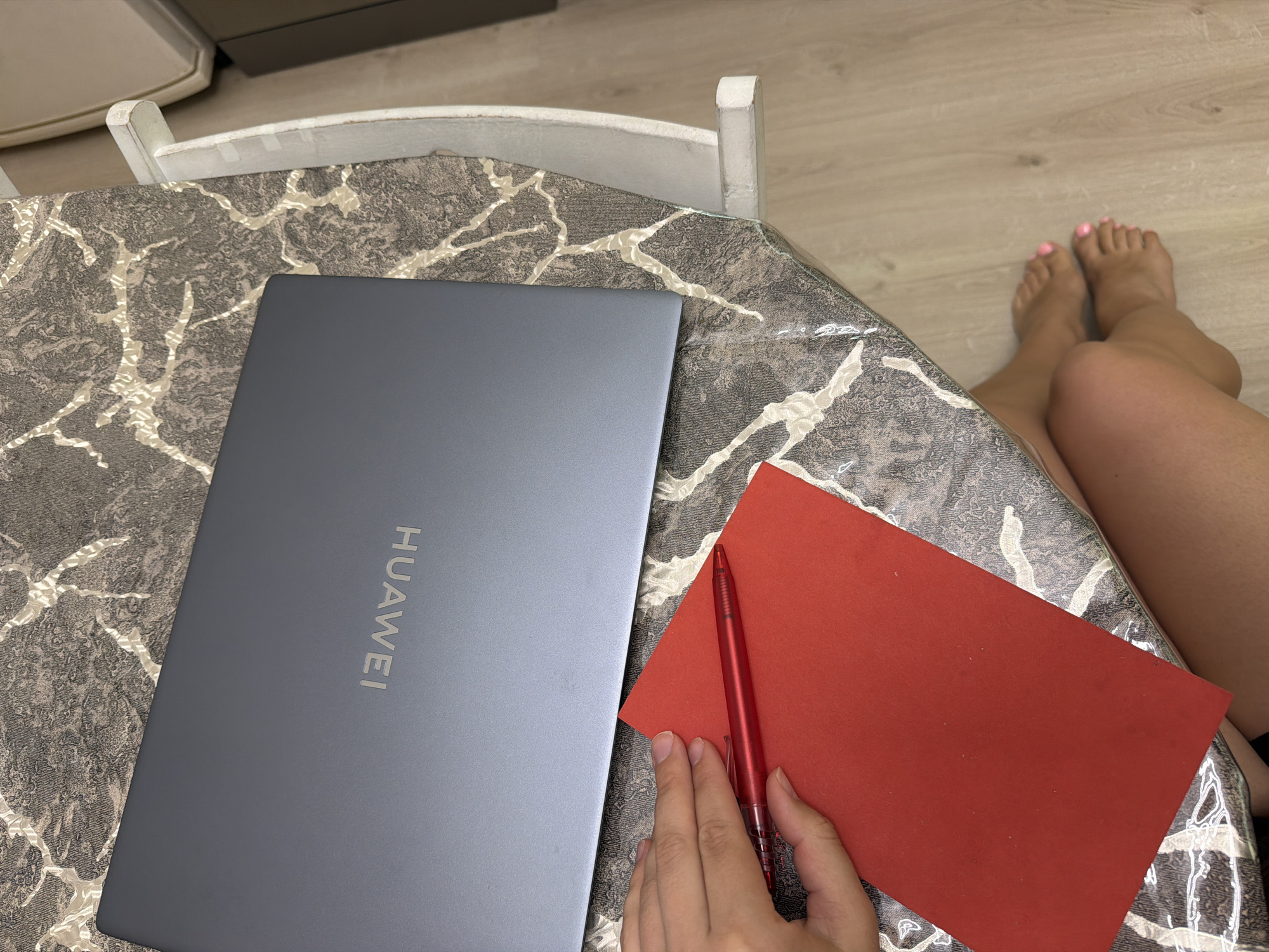 A high-angle shot shows a person seated at a table with a grey Huawei laptop, a red piece of paper, and a red pen. The table has a textured pattern in shades of grey, white, and brown. The person's legs and feet are visible, with pink nail polish on the toes. The background reveals a wooden floor. The photo does not reveal any information regarding location, especially if in Sochi, Russia.