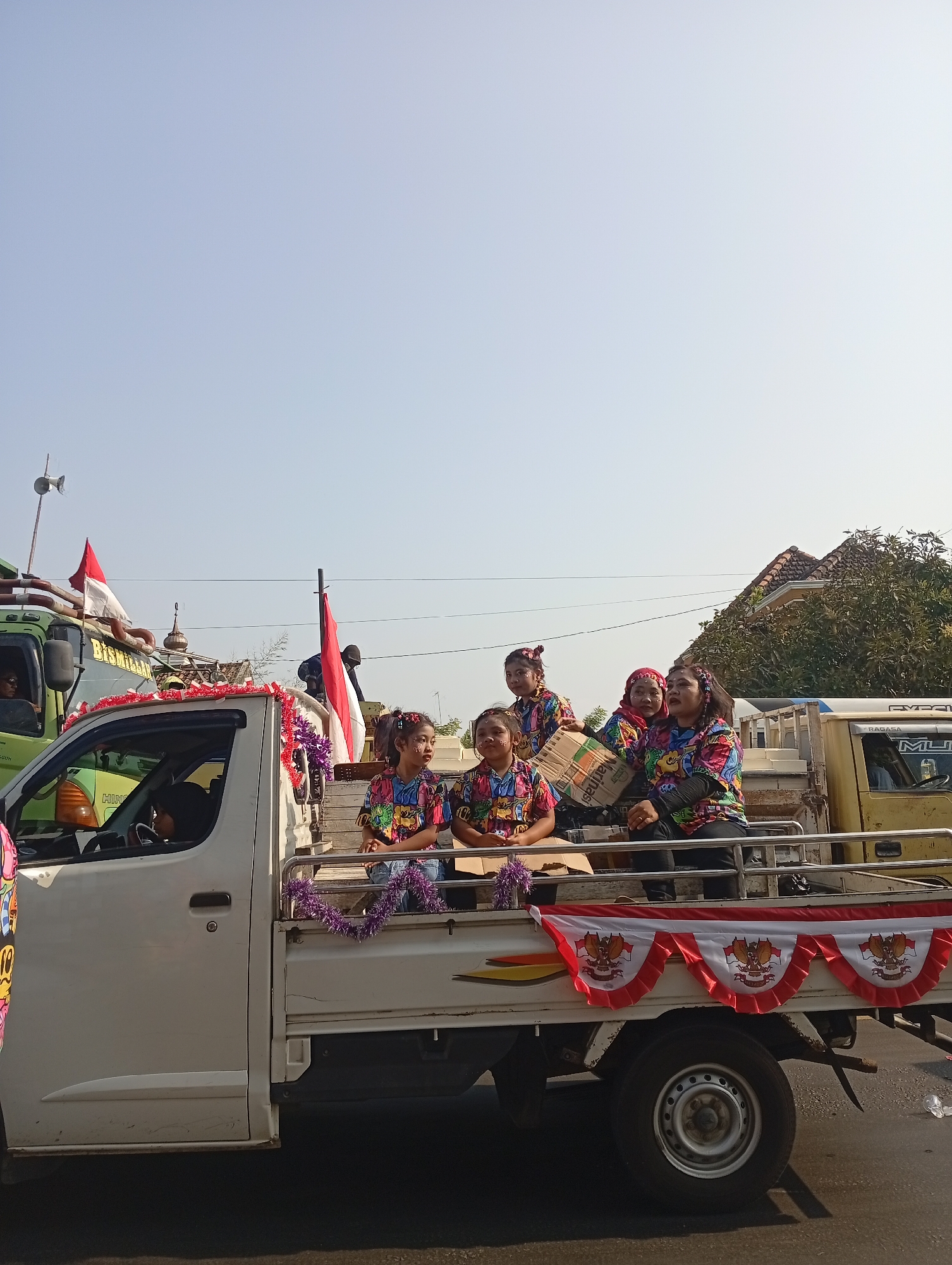 The image shows a parade in the city of Sarang, Indonesia. A white pickup truck is decorated with red and white garlands featuring the Indonesian coat of arms, and purple tinsel. Several young girls, dressed in colorful patterned shirts and with face paint, are sitting in the back of the truck. Some are holding a cardboard box. Behind them, other individuals are also visible. A green truck with the Indonesian flag is parked nearby. The scene appears to be taking place on a sunny day, with a clear blue sky overhead. Houses and trees are visible in the background.