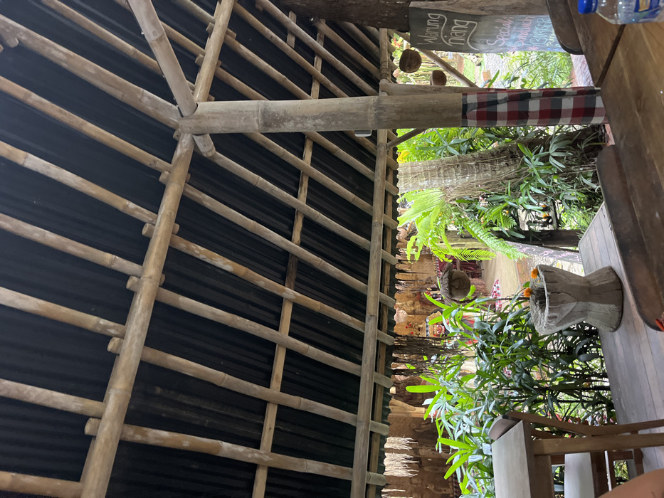 The image displays an open-air establishment in Punggul, Indonesia. The overhead structure consists of a bamboo frame supporting a dark, corrugated roof. A prominent bamboo support pillar, wrapped in a red and white checkered saput poleng cloth, stands on the right.

In the immediate foreground, a dark wooden table with a blue plastic water bottle is visible on the right. To the left, a portion of a lighter wooden table and a chair are present, with a person's forearm partially visible in the lower-left corner. Centered in the midground is a light-colored, carved stone or concrete pedestal, resembling an offering bowl, containing several orange flowers.

Beyond the foreground objects, dense tropical foliage including large ferns and various green leafy plants fills the midground. Several woven or coconut shell decorative ornaments are suspended among the plants. A dark chalkboard sign on the right displays "Warung Niang" at the top, with "Specials," "Sandwich," and "Burger" written below in white script.

The background reveals a setting with traditional Balinese architecture, featuring intricate carved stone or concrete walls with red and white color accents. A red mat or carpet is visible on the ground further in the distance. No direct human interaction is depicted beyond the implied presence of the individual in the lower-left.