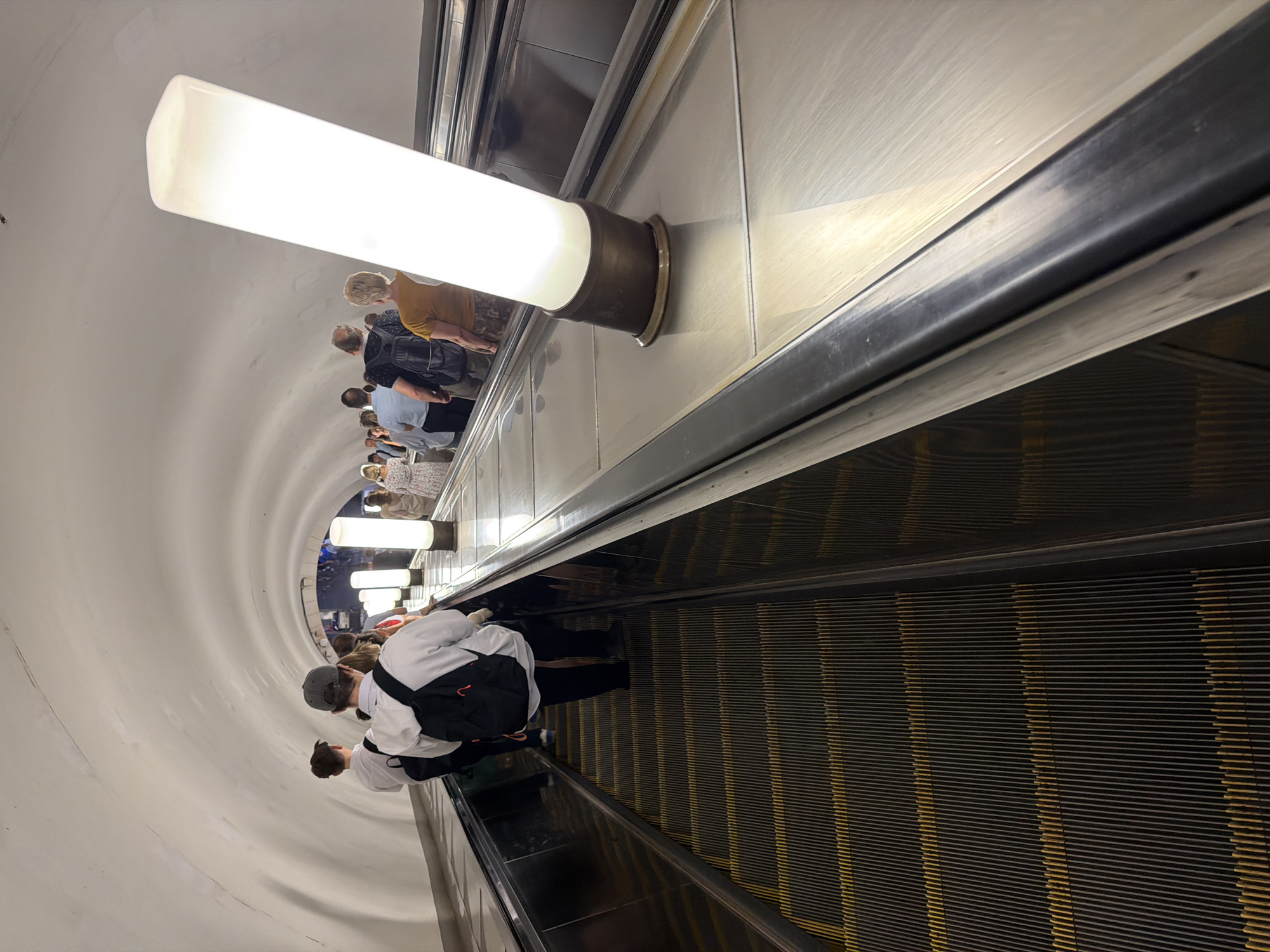 This image captures a view from an escalator in the Moscow Metro, likely in a station with a distinctive illuminated ceiling structure and tall cylindrical lights lining the escalator. A group of people are ascending the escalator, most facing forward and away from the camera. In the foreground, a person wearing a black backpack and a white hoodie stands out. To their left, another person in a white shirt is also on the escalator. Further up the escalator, more individuals are visible, some with backpacks and others with bags. The escalator itself features yellow lines on its dark, ribbed steps. The overall environment is dimly lit, with the bright lights of the escalator and the overhead illumination creating a somewhat surreal atmosphere.