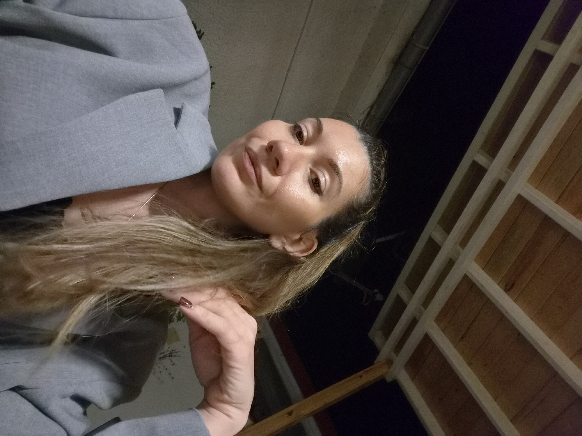 A woman is photographed from a low angle, appearing to be outside at night. She has light brown hair, fair skin, and is wearing a gray blazer. Her hand is near her face, and she is looking towards the camera. In the background, there is a partially visible structure that looks like a wooden gazebo or pergola with a dark backdrop, suggesting it is night. The lighting is dim, making it difficult to discern detailed features of the surrounding environment. The exact location in Plovdiv, Bulgaria is not identifiable from the image.