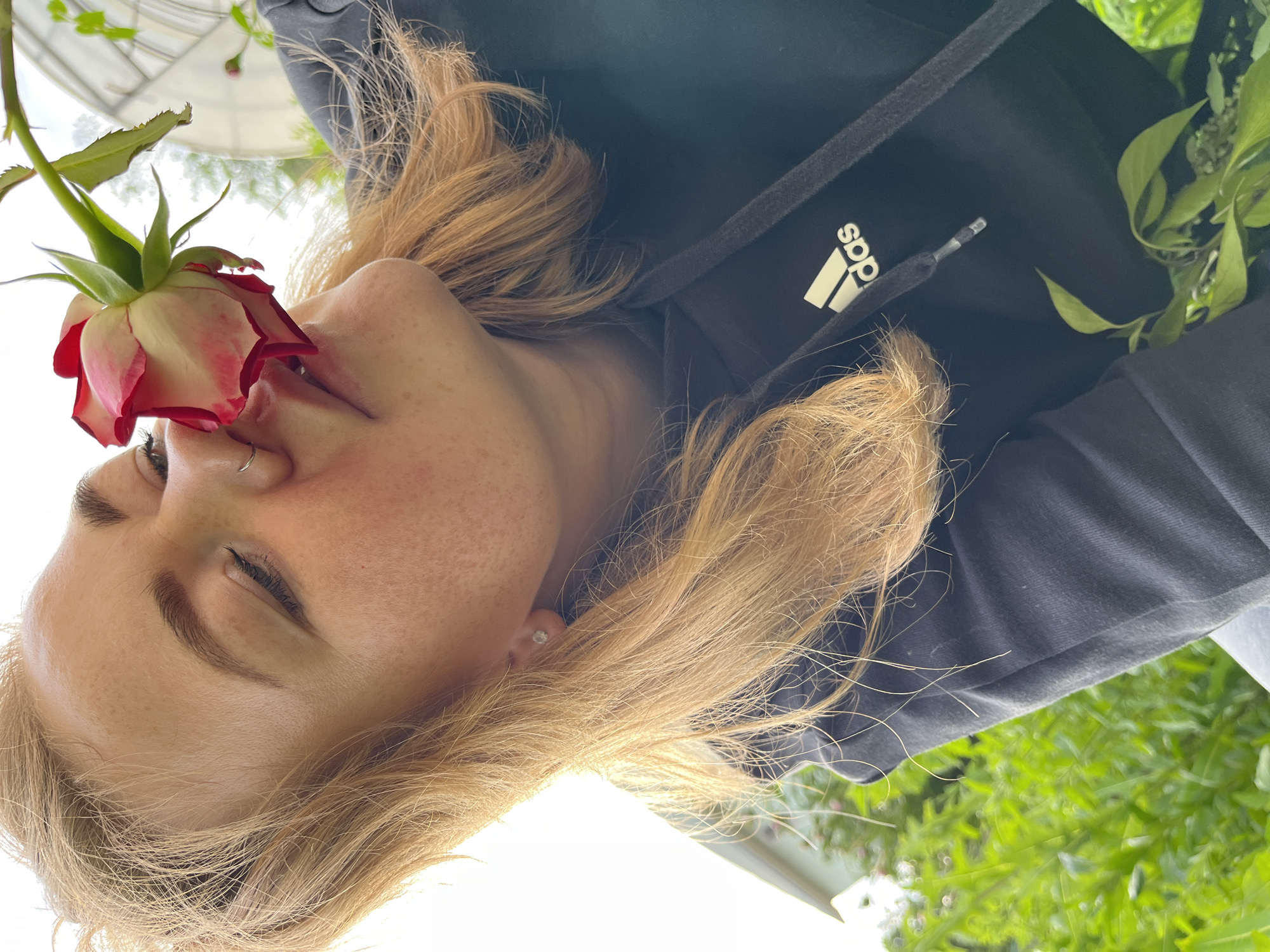 A close-up, low-angle shot captures a fair-skinned woman with blonde hair and a nose ring smelling a pink and white rose. She is wearing a black hooded sweatshirt with the Adidas logo visible. The background is a blur of green foliage and a greenhouse structure, suggesting an outdoor garden setting, possibly in or near Olkhovka, Russia. The lighting suggests it is daytime, with bright sunlight illuminating the scene.
