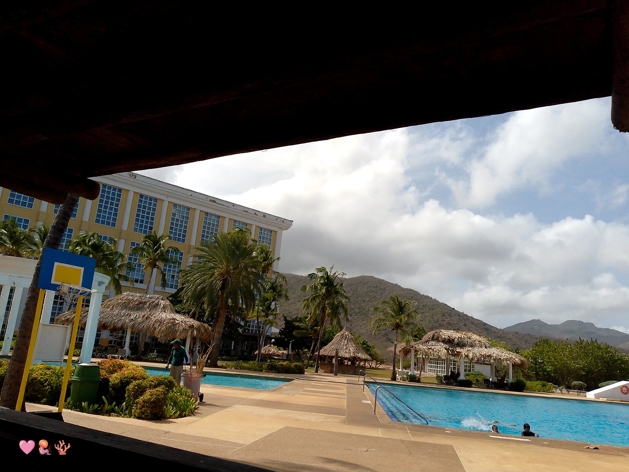 The image shows a resort or hotel in Pedro González, Venezuela. The foreground is obscured by a dark wooden structure, likely a roof or overhang. Beyond this, the scene opens to a sunny day with a cloudy sky.
To the left, a tall building with many windows can be seen. Closer, there is a basketball hoop with a yellow pole and blue backboard. A man is seen tending to the landscaping near a thatched-roof hut.
Two swimming pools are present, separated by a walkway. Two people are swimming in the pool on the right. In the background, there are mountains and more thatched-roof structures. Palm trees are prevalent throughout the scene.