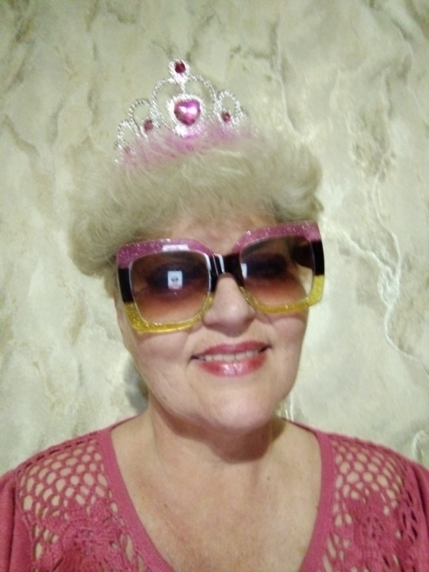 A woman with light blonde, styled hair is wearing large, multi-colored sunglasses and a silver tiara with pink accents. She is smiling, with visible lipstick, and wearing a pink crochet-like top. The background is a textured, light-colored wall. The location is potentially in Horishni Plavni, Ukraine, but there are no identifying factors visible in the image to confirm this location.