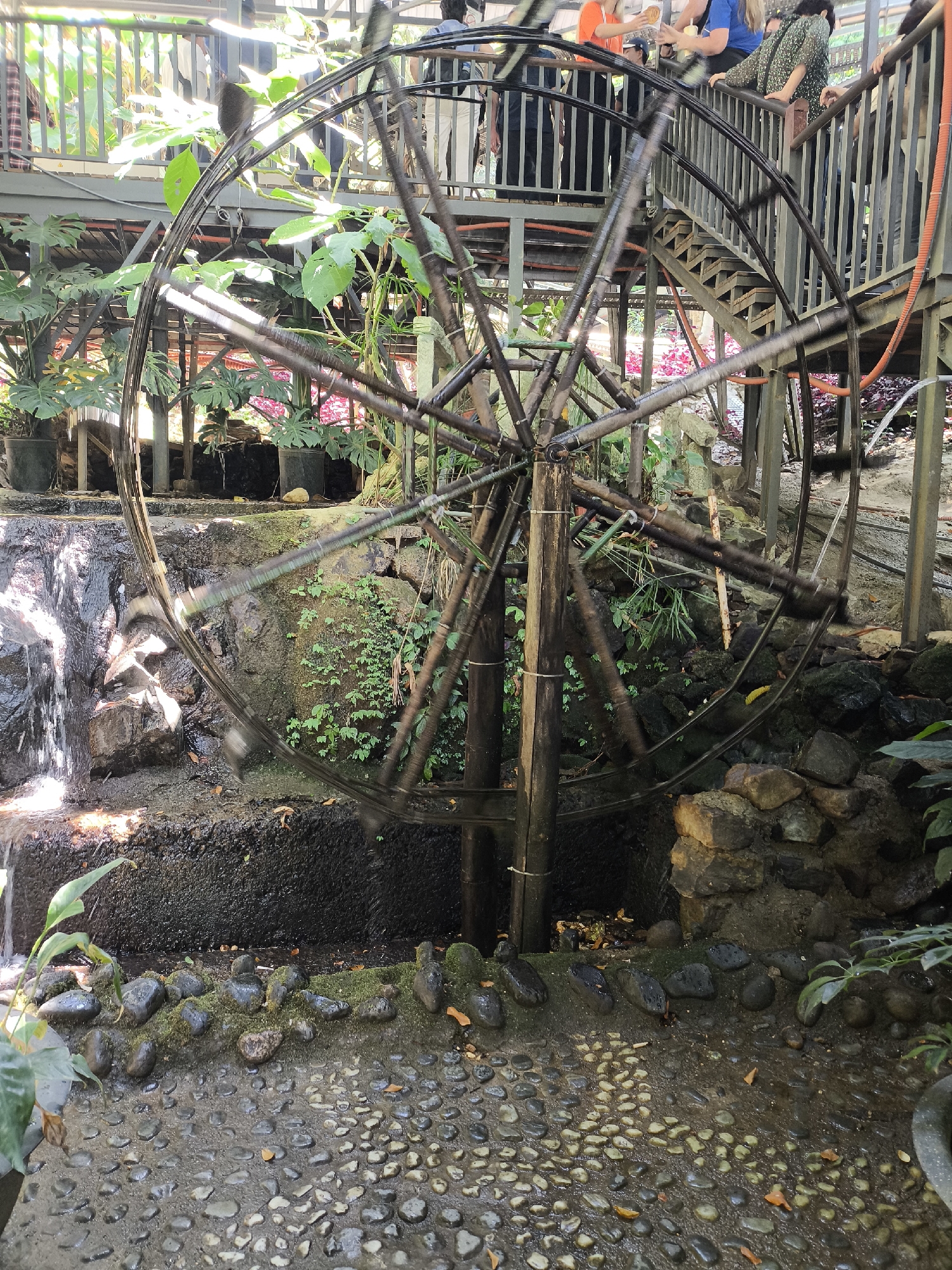 The image depicts a water wheel in motion, likely part of a decorative or functional water feature. The wheel is constructed of dark metal with what appears to be bamboo for paddles. It is located in an outdoor setting with lush green plants and rocks. Water flows into the wheel from a small, man-made waterfall. The ground is covered with small, wet pebbles. Above the water wheel, people stand on a metal platform with a metal railing. The location is Da Lat, Vietnam.