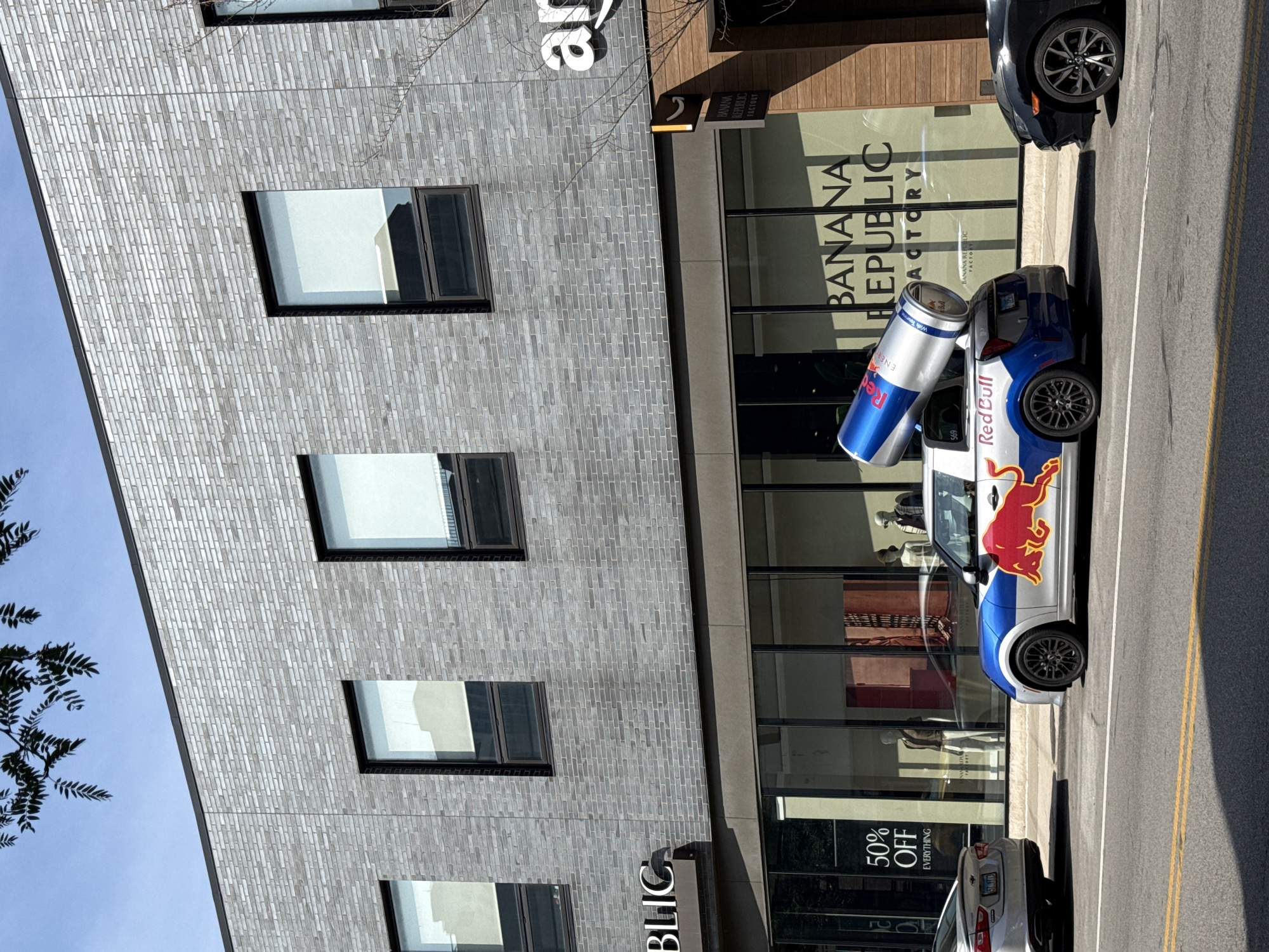 A silver Mini Cooper, wrapped with Red Bull branding and featuring a large Red Bull can on its roof, is parked on a street in front of a building with glass windows. The building has a brick facade and displays signs for "Banana Republic Factory" and "Apple." A silver sedan is parked to the left of the Mini Cooper, and a black car is parked to its right. The street is paved with asphalt and has yellow lane markings. The sky is clear and blue. The scene appears to be in a commercial area of a city.