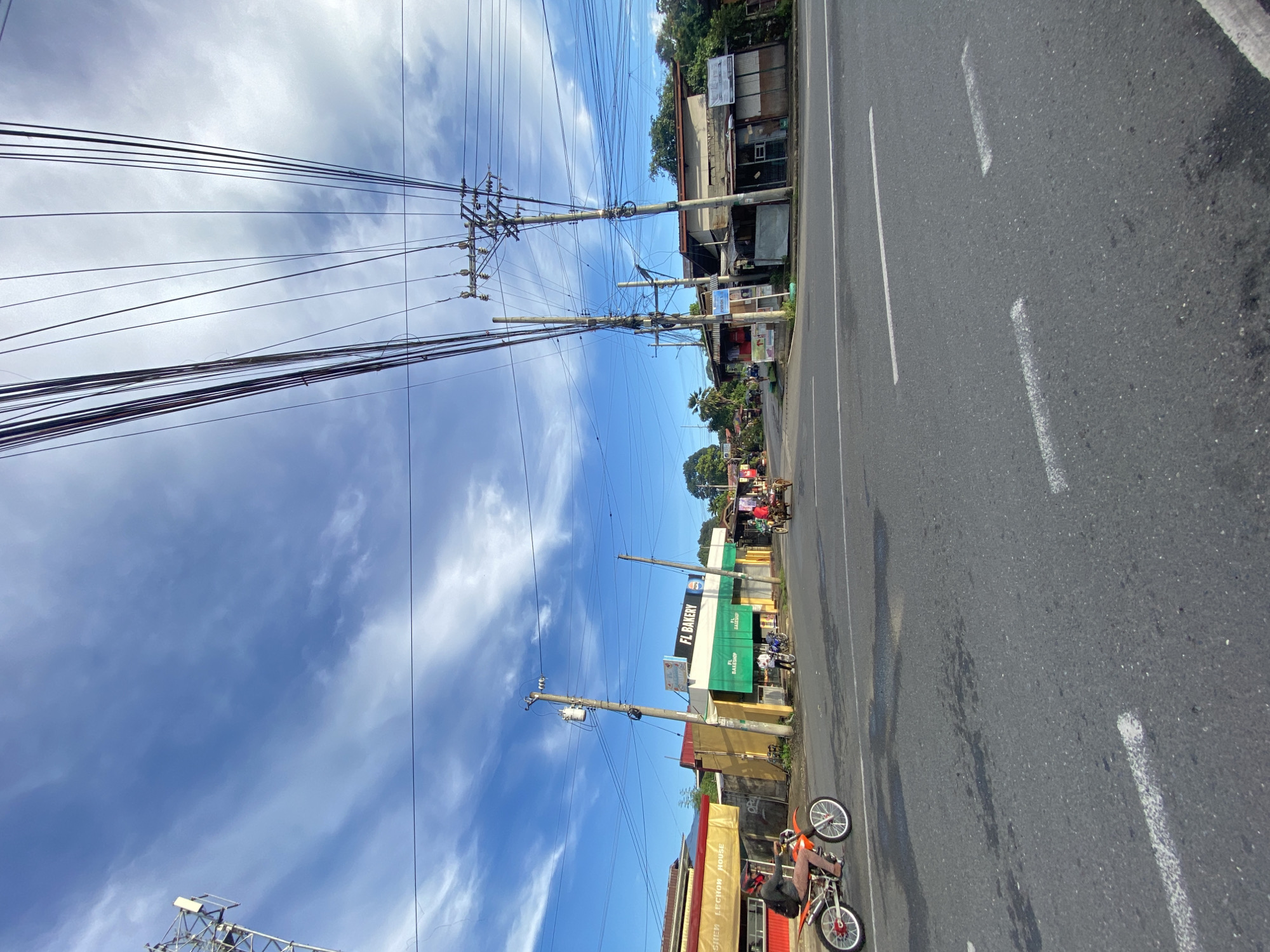 The image depicts a street scene in Davao City, Philippines, under a partly cloudy blue sky. The foreground shows an asphalt road with visible white dashed lane markings. On the left side of the road, a person is riding an orange dirt bike, wearing a helmet and casual clothing. Beside the road, there are several buildings, including a yellow and green establishment labeled "FL BAKERY." Further down the road, there are more buildings, some appearing to be shops or houses, and a utility pole with numerous electrical wires stretching across the frame. In the distance, a horse-drawn carriage is visible, along with other vehicles and trees lining the street. The environment suggests a typical urban or semi-urban setting in the Philippines, characterized by dense overhead wiring and local businesses.