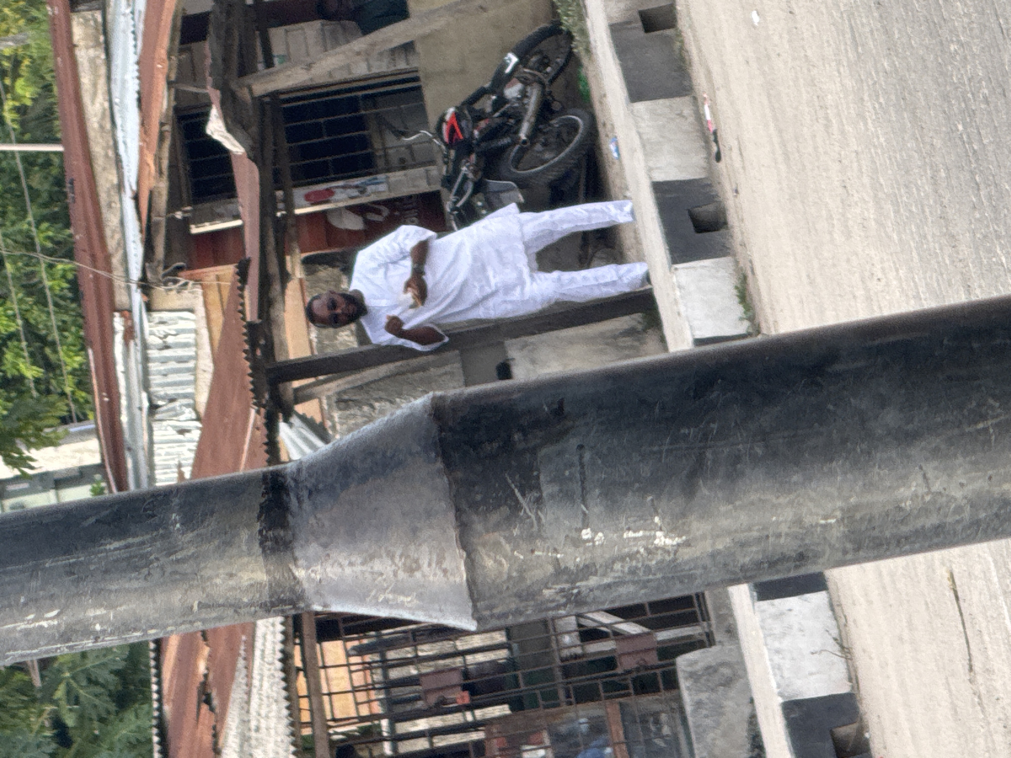 The image shows an area in Badagry, Nigeria, with a man dressed in white traditional attire standing near a motorcycle parked along a concrete pavement. The surrounding area includes buildings with corrugated iron roofs and barred windows. A thick, cylindrical, black pipe dominates the foreground.