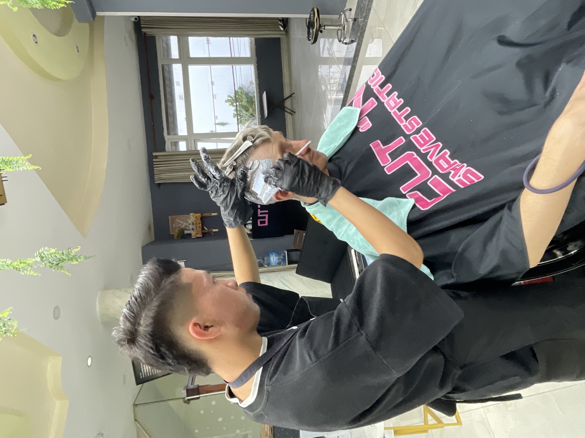 A person with short, dark, spiky hair is seen applying a light-colored substance to the hair of another person, who has short, blonde, wavy hair, using a comb and gloved hands. The person with dark hair wears a black t-shirt and a black apron. The person with blonde hair wears a black cape with pink lettering that says "CUT & SHAVE STATION." The scene appears to be a barbershop or hair salon, with polished floors, grey walls, and large windows in the background. Decorative hanging plants are visible on the ceiling. The overall environment is clean and well-lit.