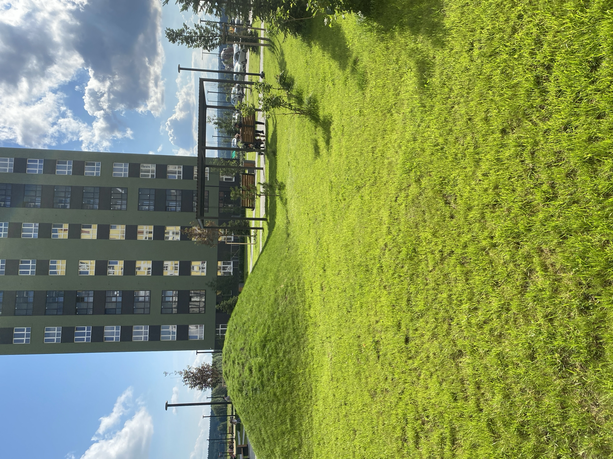 An outdoor, wide-angle shot captures a modern, green residential building and a manicured grassy area. The building, characterized by its dark-framed windows, rises tall against a blue sky dotted with white clouds. In the foreground, a rounded grassy hill slopes gently towards the viewer, covered in vibrant green grass. To the right of the hill, a sheltered seating area with benches and swings is visible, with a few people seated. The background shows more trees, a parking lot with cars, and a distant treeline, suggesting a suburban or park-like setting within a city. The scene appears to be in Yekaterinburg, Russia, based on the context provided.
