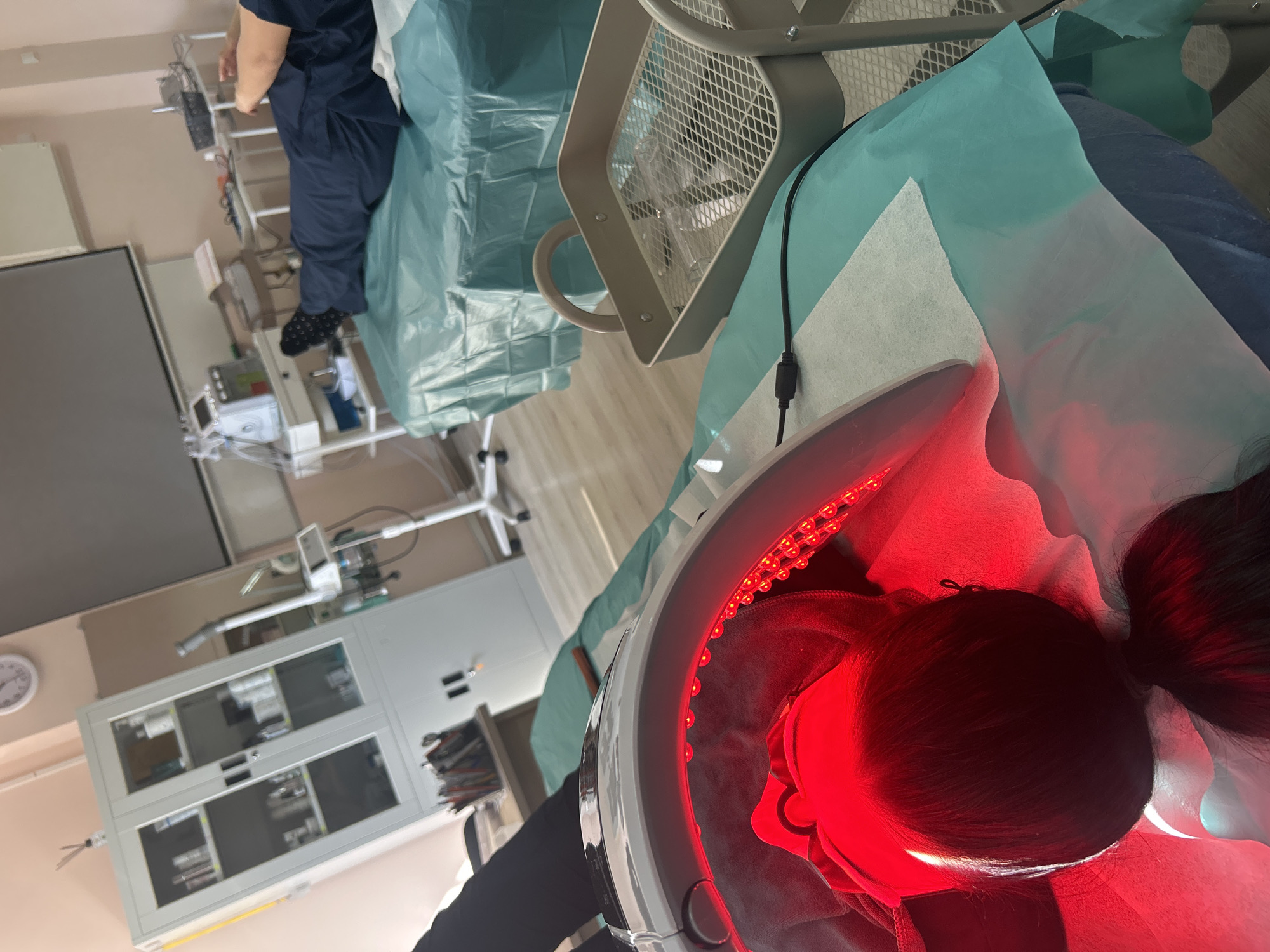 The photograph depicts an individual undergoing a red light therapy session. The subject's head is positioned under a dome-shaped device emitting red light. The person is lying on a medical examination table covered with a blue-green surgical drape and a white absorbent pad. A medical professional in blue scrubs is visible in the background, near medical equipment. The environment appears to be a medical or aesthetic treatment room, featuring a cabinet with glass doors, medical machinery, and overhead lighting. The location is unspecified.