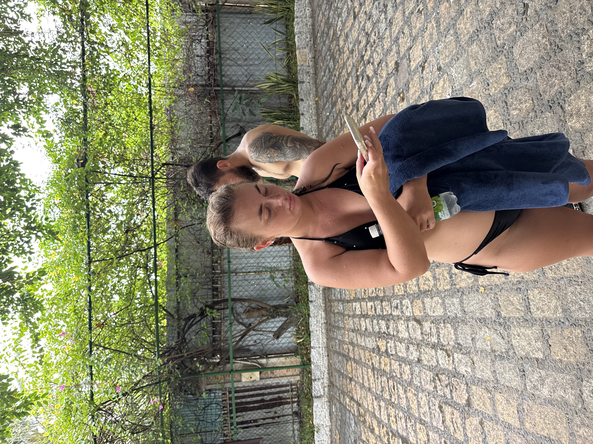 In Nha Trang, Vietnam, a woman wearing a black bikini stands on a cobblestone patio, looking at her phone. She is holding a dark blue towel and a plastic bottle of water. Behind her, a man with a beard and tattoos on his arm stands partially obscured by foliage. The background shows a fence and lush green trees.