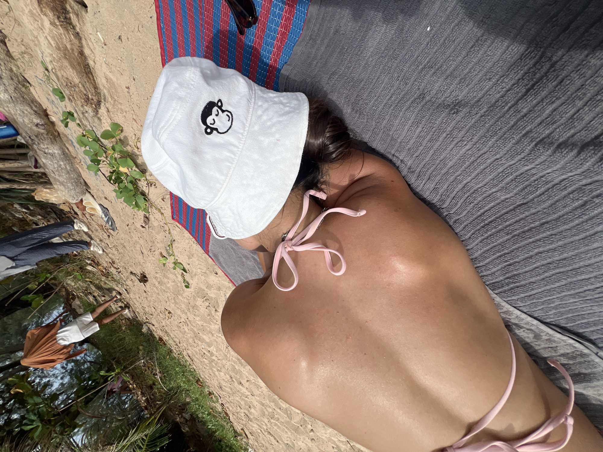 The image depicts a beach scene in Choeng Thale, Thailand. A person is lying on their stomach on a grey towel placed on a striped mat. They are wearing a pink bikini and a white bucket hat with a monkey design. Their back is visible, showing sun-kissed skin. In the background, other individuals are standing near some trees and logs on the sandy shore. The environment appears to be a tropical beach with vegetation and driftwood.