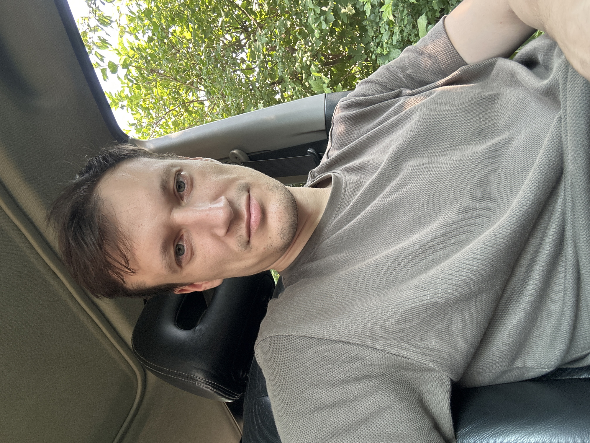 A man is sitting in the driver's seat of a car. He is looking directly at the camera. The car's sunroof is open, and trees are visible outside the car window. The man is wearing a grey t-shirt. The background is a blurry green of foliage. There are no other people or objects visible in the image. The location of Shatava, Ukraine is not depicted in this image.