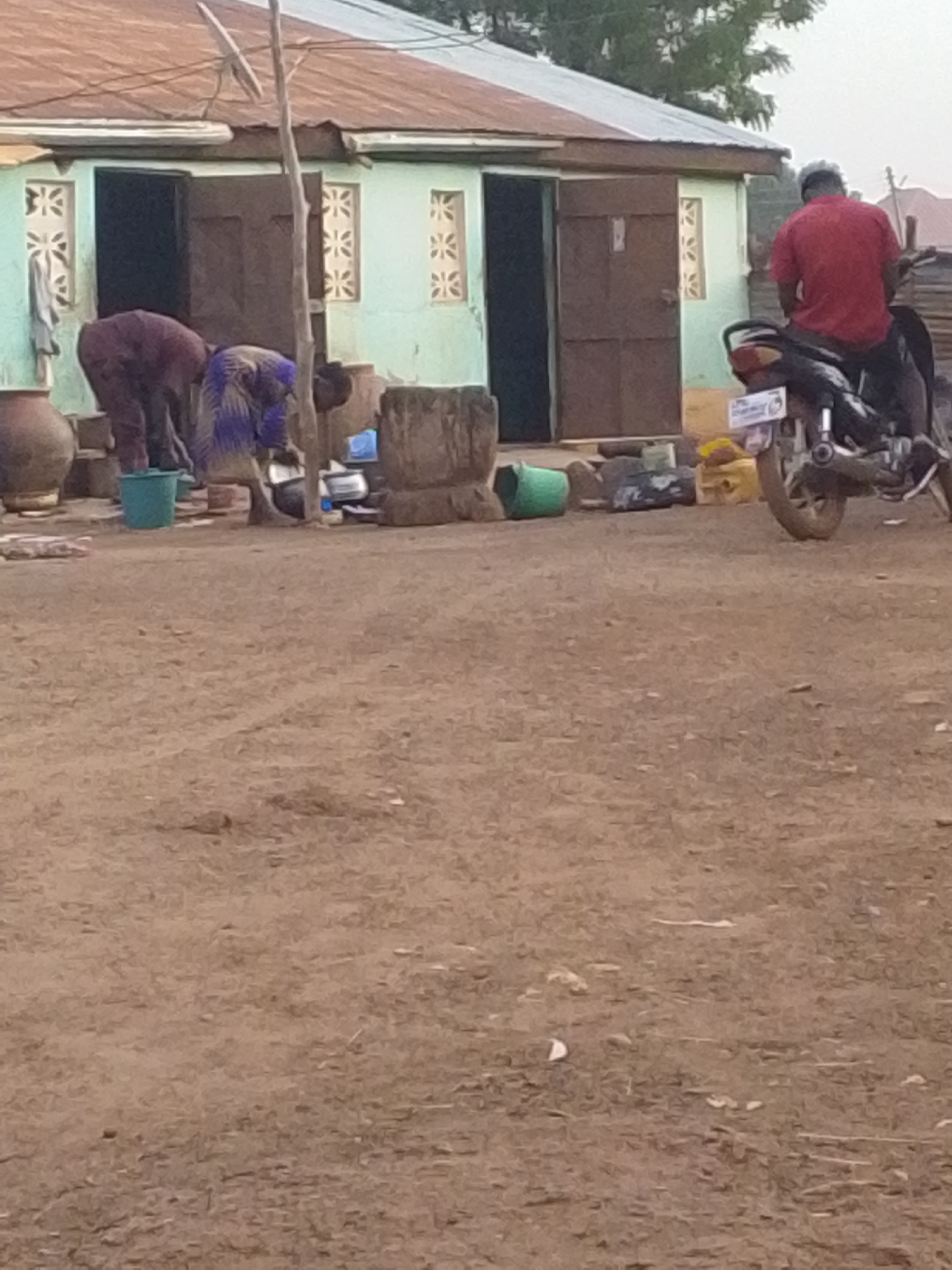 In Tamale, Ghana, the scene depicts a modest, light-green building with dark brown doors. Two women are engaged in food preparation outside the house, near a collection of pots, buckets, and cooking utensils. A man is sitting on a motorcycle. The setting is a dirt yard with some sparse vegetation.
