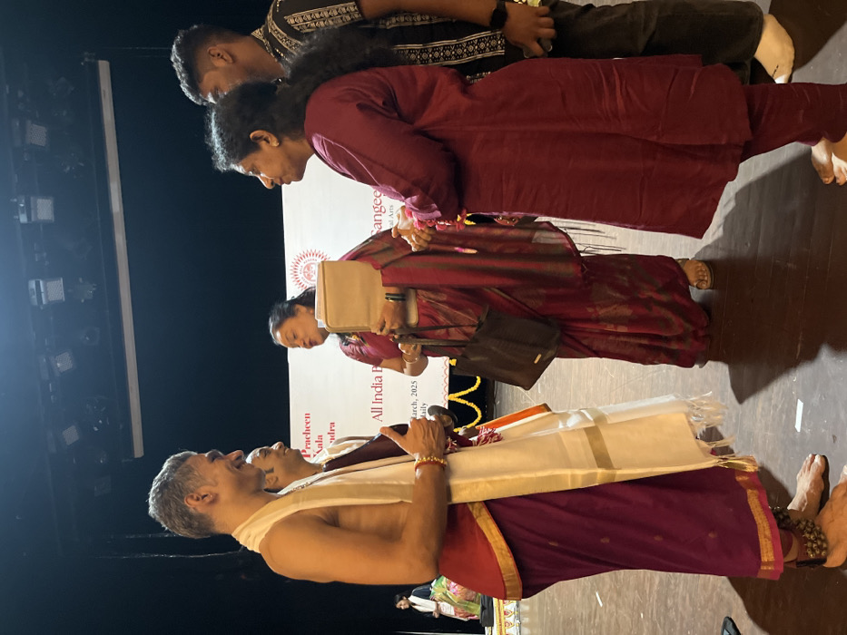 The scene takes place on a stage in the city of Chandigarh, India, at the coordinates (30.733, 76.79). There are four individuals on stage. Two are wearing traditional Indian clothing, one is wearing a red dress and another is wearing a long maroon garment. The individual in the red dress is holding a binder, and the individual in the long maroon garment is standing with hands clasped in front of them. The two other individuals are standing in front of a banner that reads "Pracheen Kala Kendra, All India Festival of Traditional Arts, March, 2023." The individual on the left is wearing a white dhoti and a dark maroon garment. They are standing with their right hand raised in a thumbs-up gesture, while the individual in the long maroon garment looks to be speaking. The floor is a dark wooden floor with dark brown accents. There are spotlights above and to the right of the stage, as well as a dark background with a large rectangular screen.