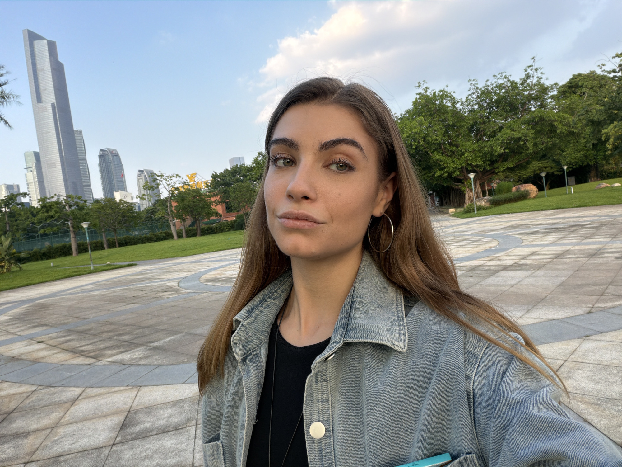 A young woman with long brown hair is taking a selfie in a park. She is wearing a denim jacket and a black shirt, and large hoop earrings. The park has a paved walkway with circular patterns, green grass, and trees. In the background, several modern skyscrapers are visible against a blue sky with clouds. The scene appears to be in the Yuexiu District of Guangzhou, China, a common area for such mixed urban and green spaces.