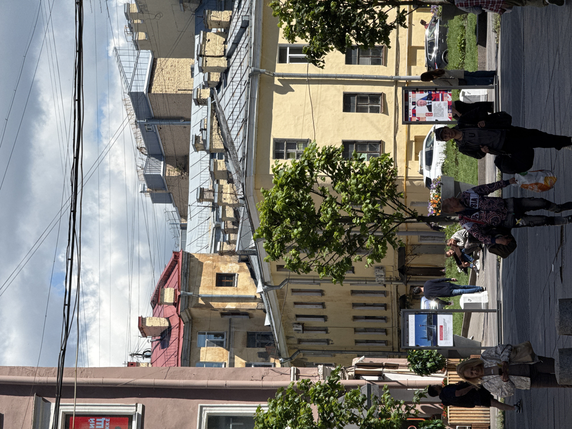 This image captures a street scene in Saint Petersburg, Russia, likely during daylight hours given the strong shadows. In the foreground, several individuals are walking across a paved area, suggesting a public thoroughfare. A woman in a light-colored jacket is looking at her phone, and two men are walking towards the viewer, one carrying a bag and a plastic bag filled with items, the other in a patterned jacket.
In the middle ground, more people are present. Some are seated on a grassy area, while others stand or walk. One man in a dark sweater and jeans is walking away from the camera. A large display board shows an image of a stadium and text, possibly advertising an event or attraction. Another display board features a portrait of a man in a suit, along with text and a geometric pattern.
The background is dominated by multi-story buildings with distinct architectural styles, characteristic of Saint Petersburg. The buildings are painted in shades of yellow and pink, with red tiled roofs and numerous windows. Overhead, a network of electrical wires crisscrosses the sky, a common feature in urban environments. Trees with green foliage are visible on either side of the street, adding a touch of nature to the cityscape. Several cars are parked or driving in the distance, indicating an active urban setting. The sky is partly cloudy with patches of blue sky visible.