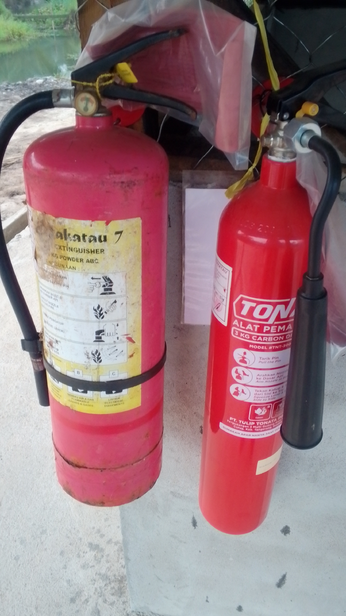 Two red fire extinguishers are positioned outdoors on a concrete surface. The fire extinguisher on the left, a larger model, is labeled "akatau 7" and appears to be a dry powder ABC type. It has a black hose and a pressure gauge. The fire extinguisher on the right is a smaller model, labeled "TON" and "3 KG CARBON DI". It also has a black hose and a pressure valve. Both extinguishers are secured with straps and are partially covered by plastic sheeting in the background. The environment suggests an outdoor storage area, possibly near a body of water and vegetation, which could be consistent with a location in Ambawang, Indonesia, where such equipment might be stored. The concrete surface shows some discoloration and small dark spots.