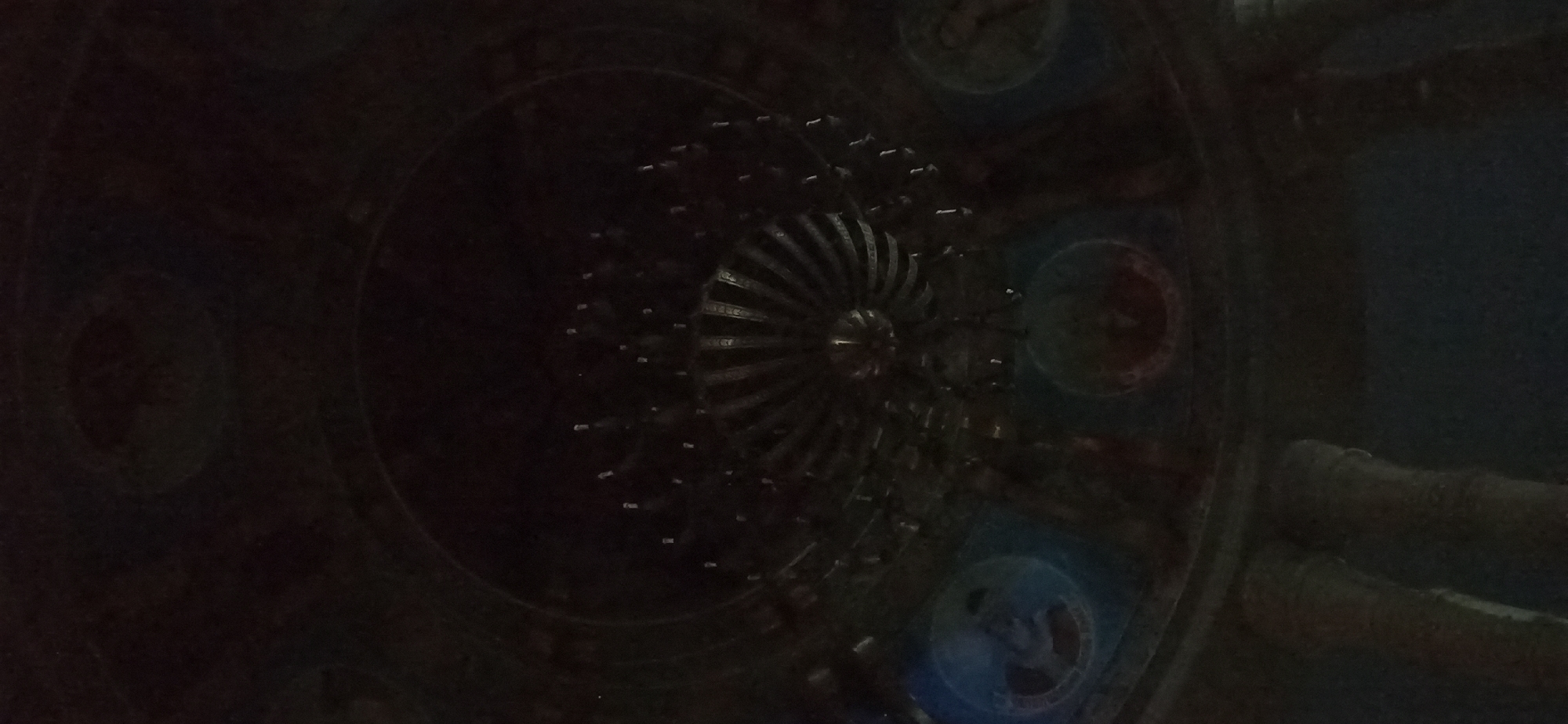 This is a dimly lit, low-angle shot of a chandelier hanging from a domed ceiling in Dushanbe, Tajikistan. The chandelier is ornate and appears to be made of metal, with multiple tiers and many small light bulbs. The ceiling is decorated with circular paintings, two of which are visible in the frame, one with blue and yellow tones and the other with red and orange tones. The paintings depict figures, though they are not clearly discernible due to the poor lighting and low resolution of the image. The overall impression is of a grand interior space, possibly a religious building or a theater, in the city of Dushanbe.
