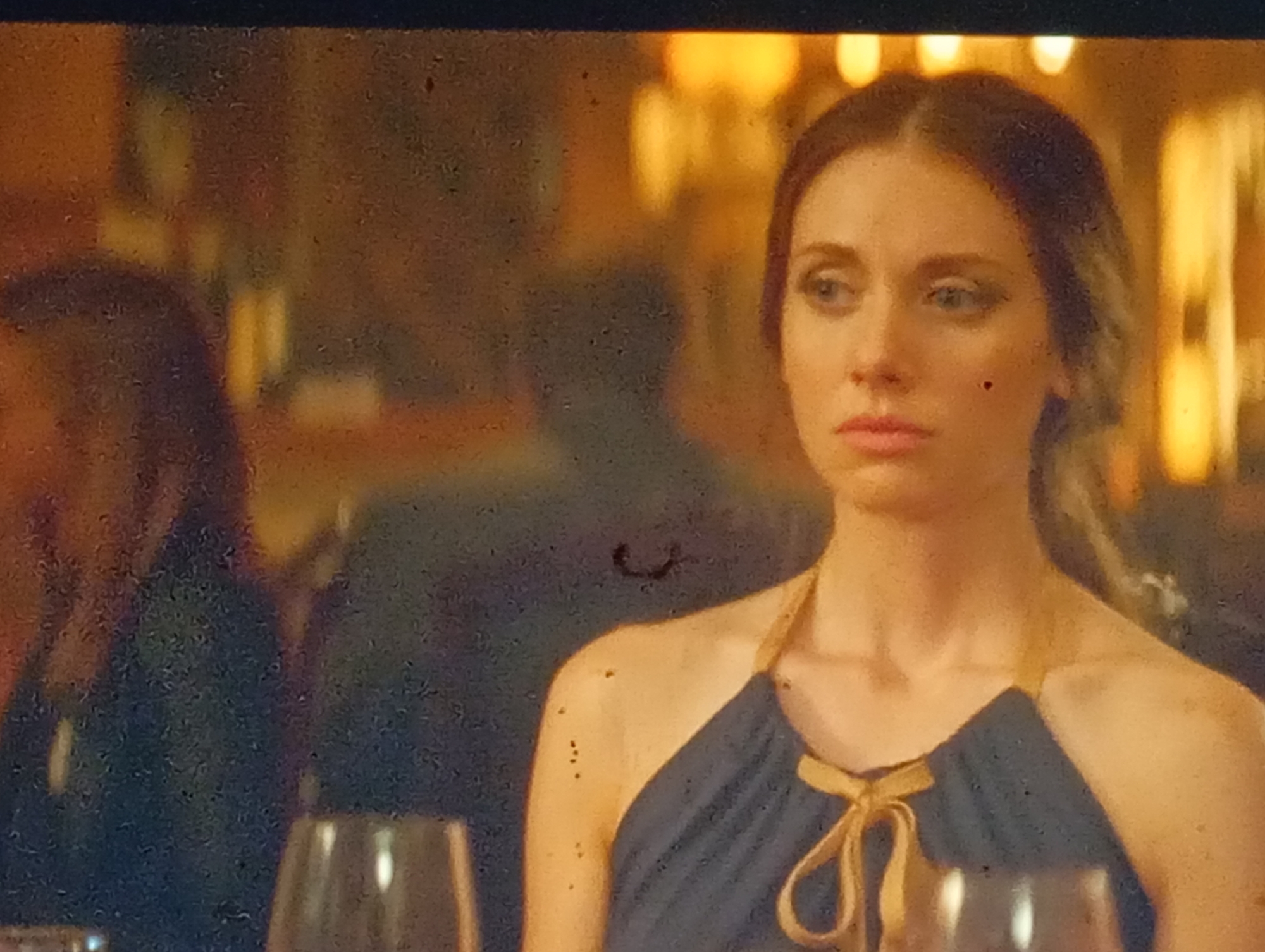 The original image is a medium shot of a woman with long, dark hair, wearing a dark blue halter dress with a yellow tie. She is looking to her left with a neutral expression. The background is blurred and appears to be a dimly lit indoor setting with warm lighting, possibly a restaurant or event space. There are reflections of light sources in the background. To the left of the woman, a portion of another person's shoulder and dark clothing is visible. In the foreground, two wine glasses are partially visible. The image quality is degraded by numerous specks and scratches, indicating it might be a photograph of a screen display. The scene does not provide any specific indicators that it is located in Sofiivska Borshchahivka, Ukraine.