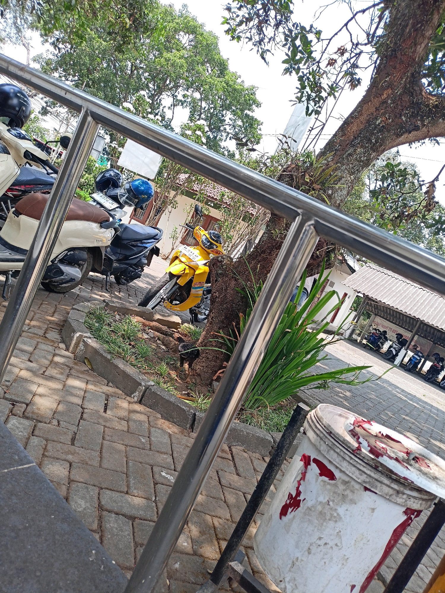 The image shows a parking area outside a building in Kewaluhan, Indonesia. Several motorcycles are parked in the foreground, some with helmets on them. A large tree with dense foliage is visible on the right side of the image, and a metal railing runs across the foreground. In the background, a covered parking structure houses more motorcycles. The ground is paved with interlocking bricks, and there is some vegetation and a trash can in the scene. The overall impression is one of an outdoor public space, possibly near a residential or commercial area.