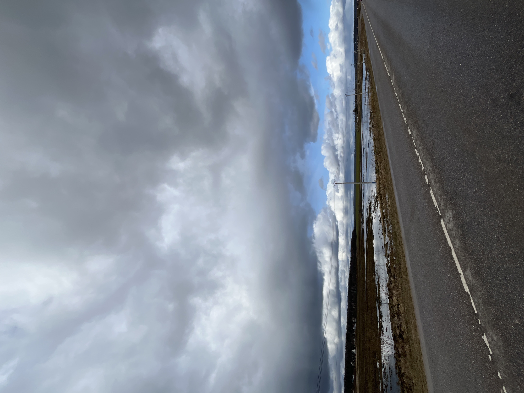 A slightly off-vertical view showcases a road scene near Ямуга, Russia. The right portion displays a dark asphalt road with a worn white line marking the edge. To the left of the road is a waterlogged field, reflecting the sky. Utility poles stand along the field's edge. The sky is dramatically divided, with heavy, dark storm clouds dominating the left and lighter, partly cloudy blue skies on the right.