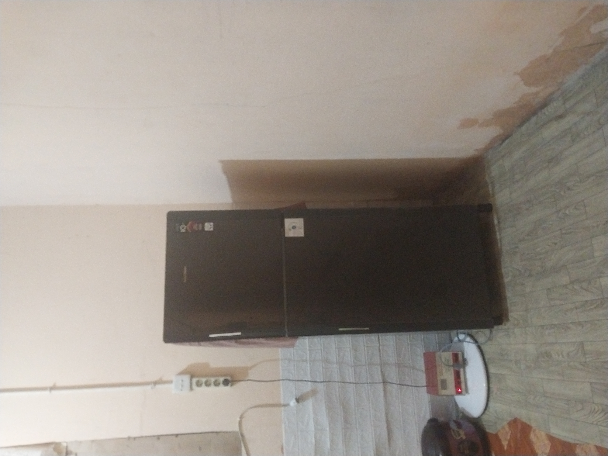 The image shows a corner of a room. On the right, a dark-colored, two-door refrigerator stands against a light-colored wall. The wall exhibits some discoloration and peeling paint near the floor. To the left of the refrigerator, a white tiled wall is visible. A white electrical outlet with multiple sockets is mounted on the beige wall above the tiled wall. A white cord extends from the outlet and runs down behind a red and white electrical device situated on a circular white mat on the light-colored wooden floor. To the left of the electrical device, part of a dark-colored object, possibly a rice cooker, is visible. The floor is covered with light-colored wooden planks. The scene appears to be an indoor domestic setting in Air Tiris, Indonesia.