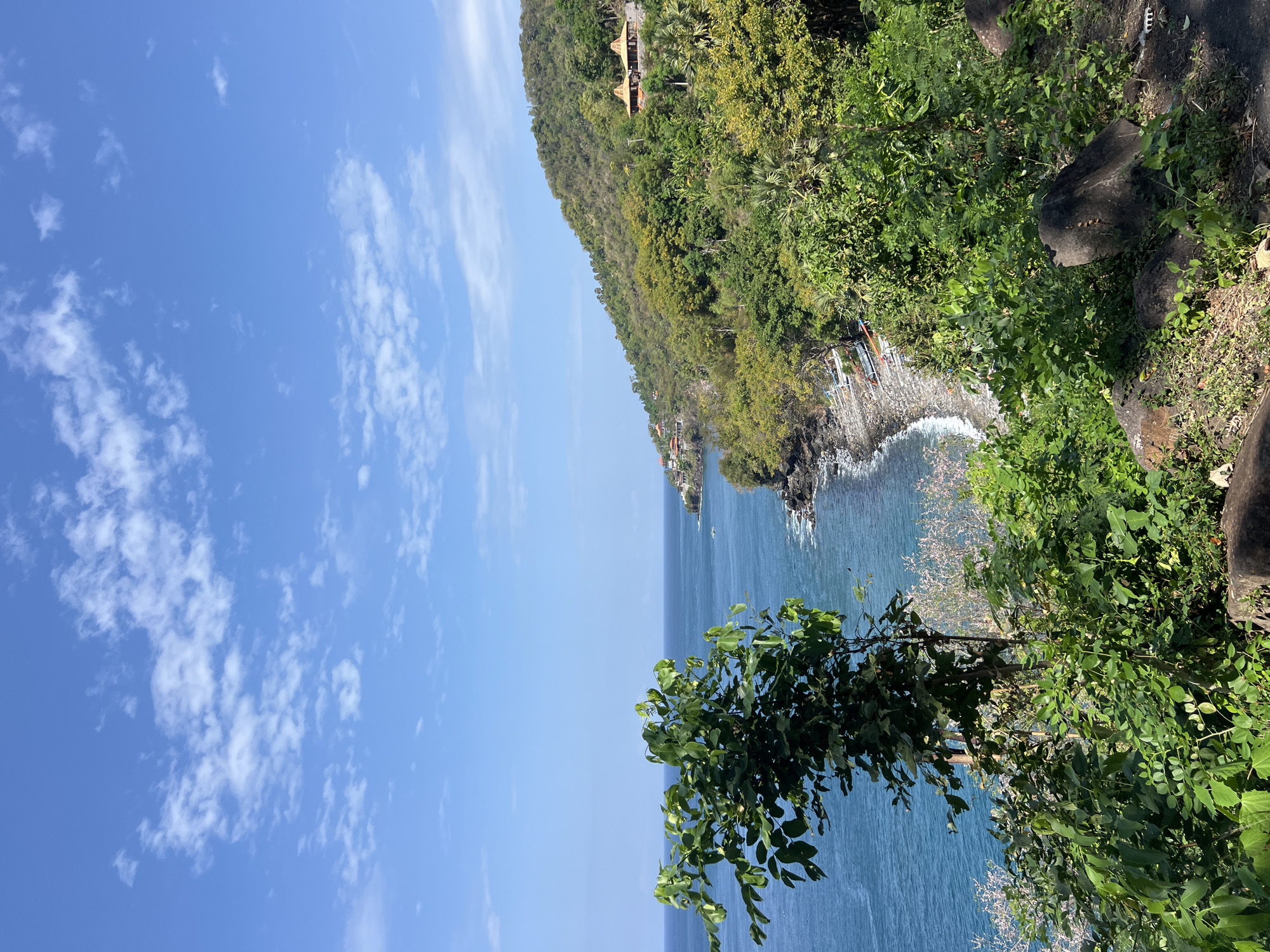 The image captures a scenic view of the coastline near Abang, Indonesia. The ocean, a vibrant shade of blue, meets a rocky shore lined with small boats. Lush green vegetation covers the rolling hills that slope down towards the sea, dotted with a few buildings that appear to be traditional Balinese structures with thatched roofs. The sky above is a clear blue, adorned with scattered white clouds, suggesting a bright, sunny day. The foreground is dominated by a large tree with green leaves, partially obscuring the view of the coastline. The overall impression is one of natural beauty and tranquility.