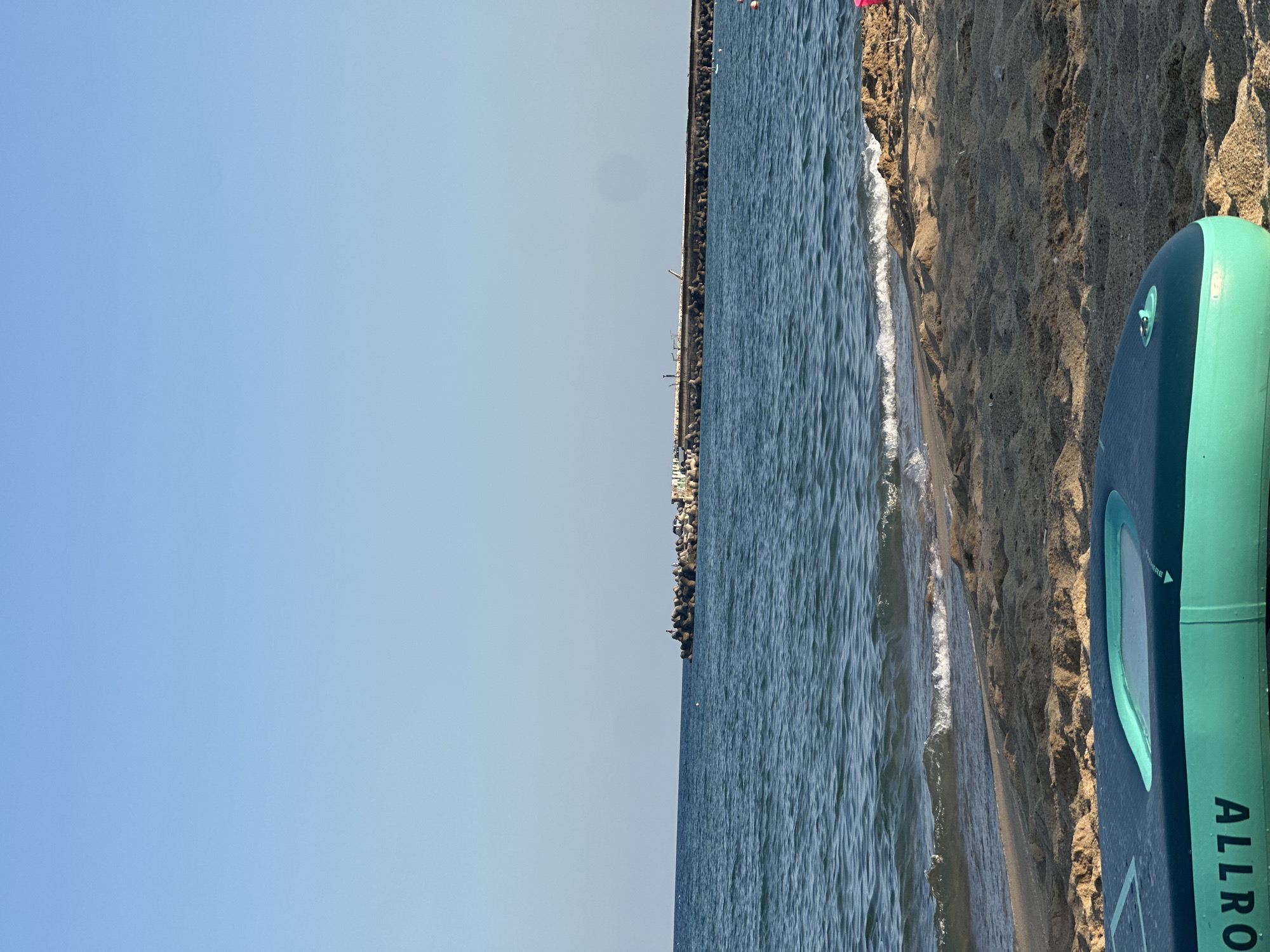 This is a media file of a beach scene. The main subjects are the ocean, the sand, and a stand-up paddleboard. The ocean is a deep blue with visible waves and a thin white foam line where it meets the shore. The sand is a light brown and appears dry and textured. Part of a dark blue and teal stand-up paddleboard is visible in the foreground on the right side of the frame. The paddleboard has a teal handle and a clear window. Text on the paddleboard reads "ALLRO..."
In the background, a long pier or jetty stretches out into the ocean, lined with a series of concrete barriers. A very distant view of structures or buildings can be seen along the coastline at the end of the pier. The sky is a clear, pale blue, indicating it is likely daytime, possibly late afternoon or early morning due to the lighting on the sand. The overall mood is serene and peaceful, suggesting a calm day at the beach. There are no people clearly visible, though the presence of a paddleboard implies recreational activity. The location is a beach, likely a coastal area with a man-made structure extending into the water.