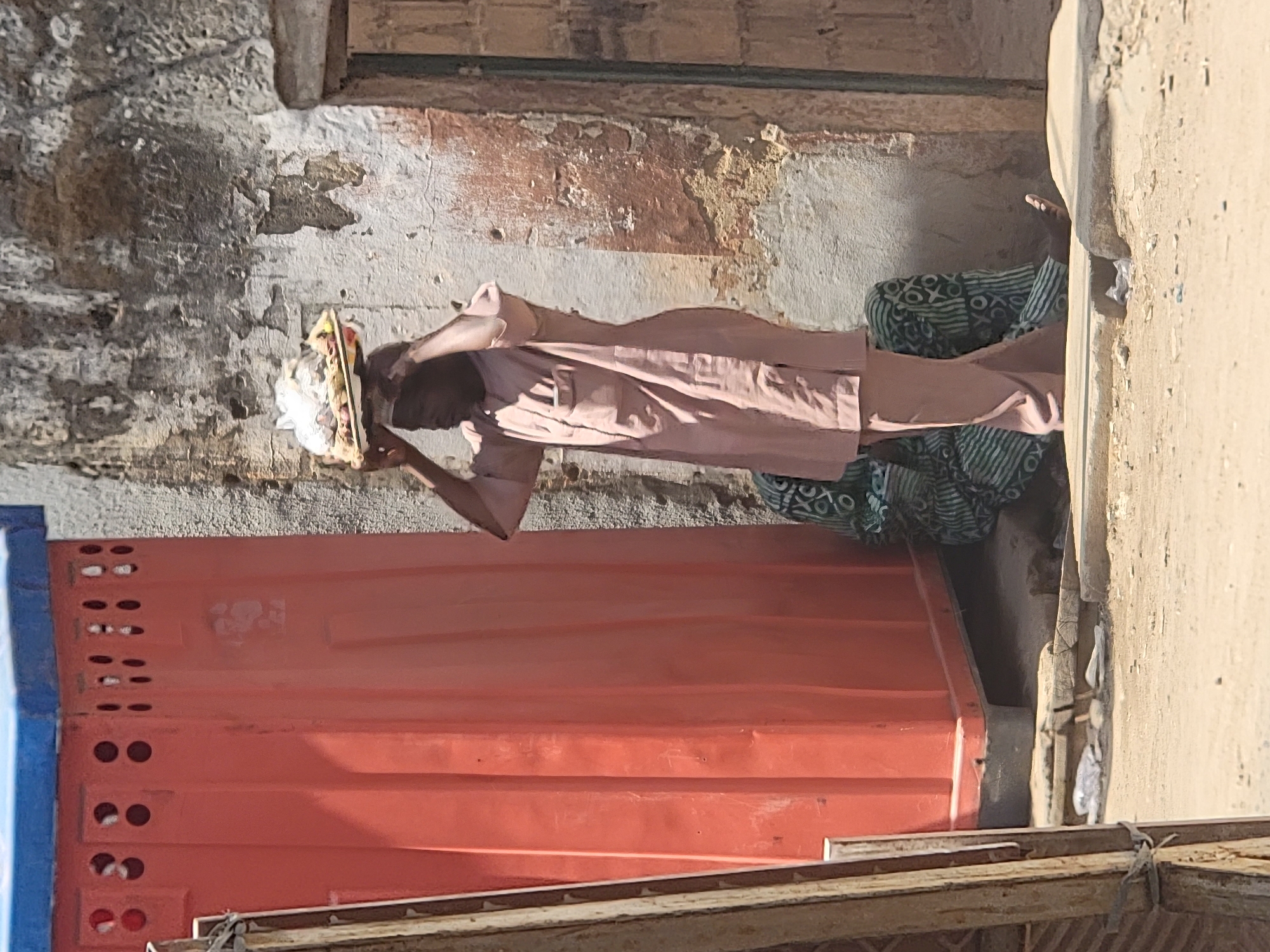 In Maiburiji, Nigeria, a man wearing a long-sleeved, button-down shirt and trousers carries a tray of white items on his head. He walks next to a low, orange container-like structure with ventilation holes and is partially covered by a green and white patterned textile on the ground. The scene is set against a backdrop of a weathered, light-colored building exterior.
