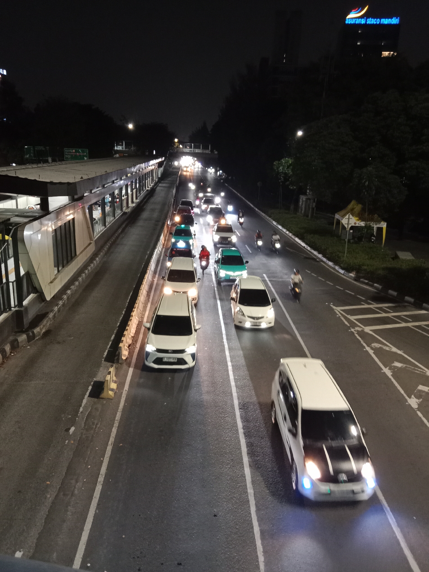 The image depicts a nighttime street scene in West Jakarta, Indonesia, characterized by moderate traffic congestion. A multi-lane road is visible, with vehicles including cars and motorcycles navigating through the flow. On the left side of the road, there is a bus stop or transit shelter with a covered waiting area. The buildings in the background are illuminated, suggesting an urban environment. The overall atmosphere is one of a busy city at night, with the headlights and taillights of vehicles creating streaks of light. The image seems to have been taken from an elevated perspective, possibly a bridge or overpass, looking down onto the road.