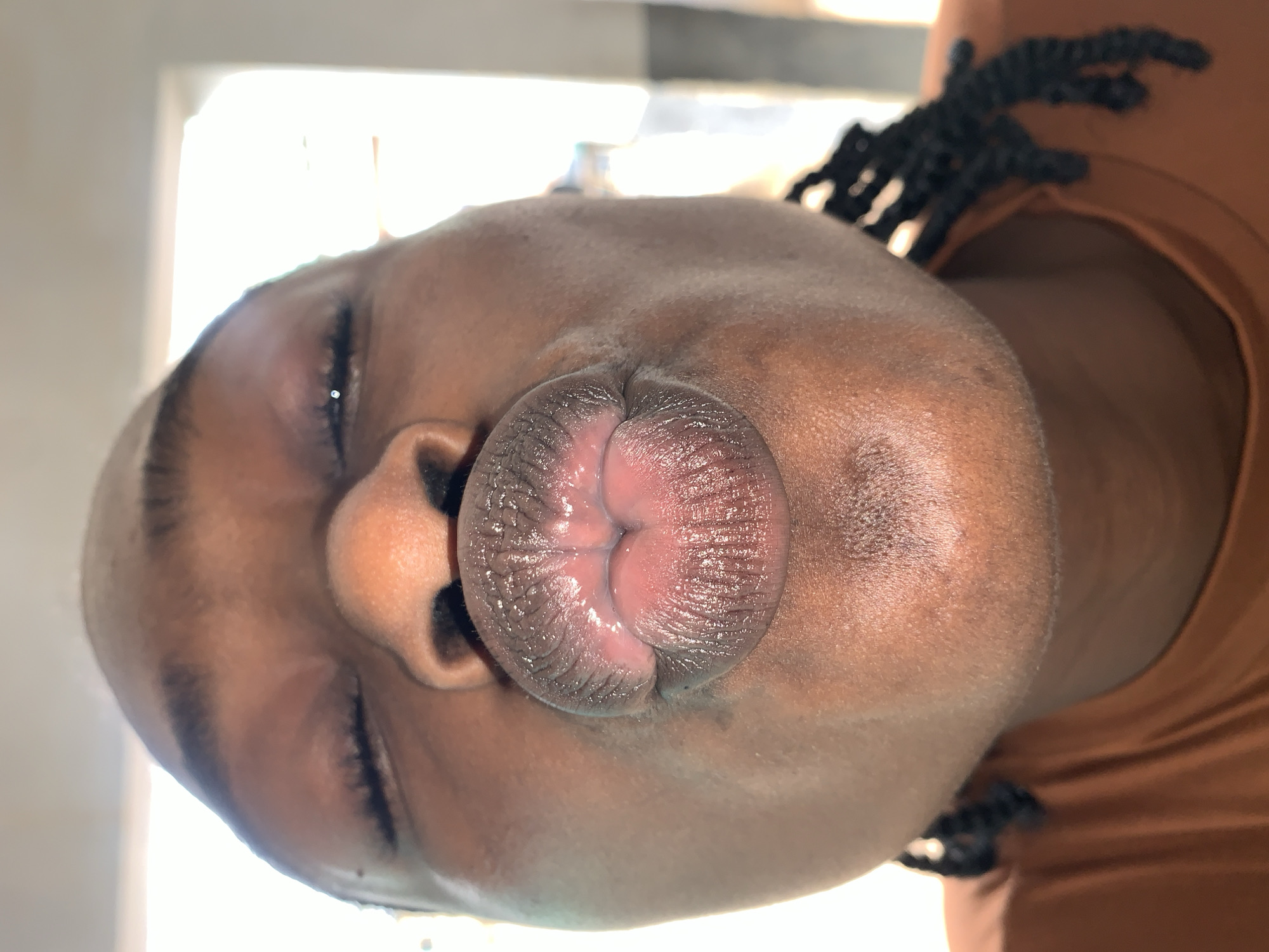 A close-up, low-angle portrait features the face of a person with dark skin, located in the city of New Stadium, Nigeria. The subject's eyes are closed, with dark eyebrows visible above them. The nose is prominent and rounded. The lips are pursed and pushed forward into a distinct pout or kissing shape, displaying visible texture and a subtle pinkish hue on the inner surfaces. Dark braids are visible near the lower right side of the frame, partially obscuring the neck and shoulder area. The subject wears an orange-brown, possibly terracotta-colored, t-shirt, visible across the lower portion of the image. The background is uniformly bright and out of focus, suggesting a strong light source or window behind the subject.