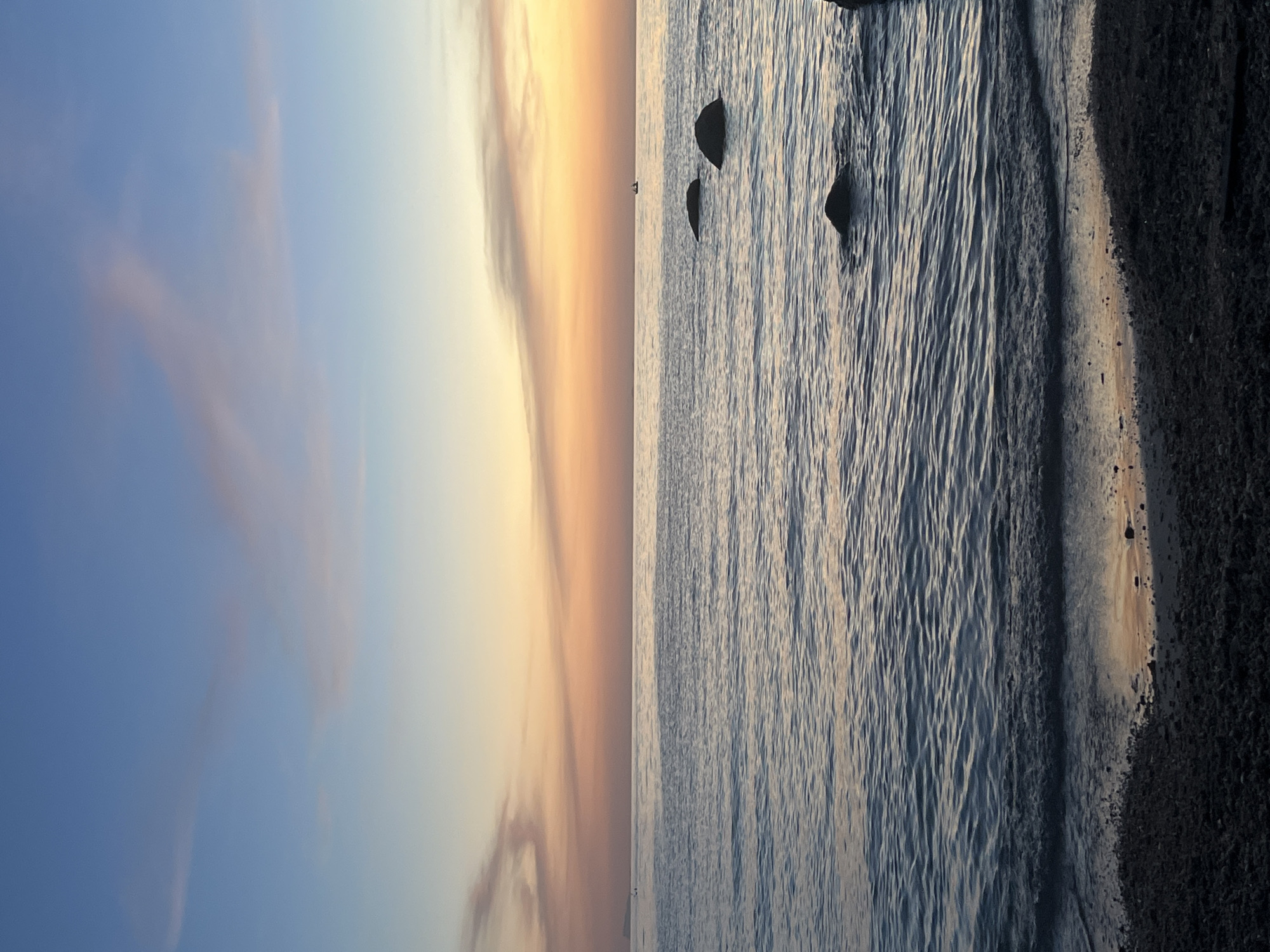 The image depicts a serene seascape at dusk, likely in Ko Pha-ngan District, Thailand, given the distinct horizon line and tropical sunset colors. The sky transitions from a pale blue at the top to warm hues of orange and pink near the horizon, with wispy clouds scattered across it. The ocean is calm, with gentle waves lapping at a pebble-strewn shore in the foreground. Several dark, rounded rocks are partially submerged in the water, adding texture to the scene. In the distance, a small boat is visible on the horizon, suggesting a quiet maritime presence. The overall atmosphere is tranquil and peaceful, capturing the beauty of a coastal sunset.