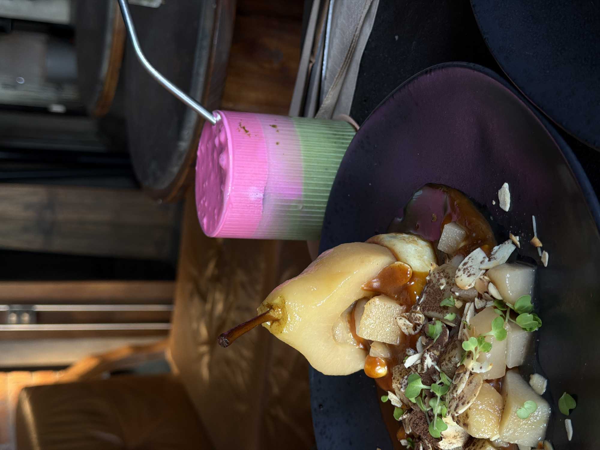 In the city of North Kuta, Indonesia, a close-up shot focuses on a dessert and a colorful drink, likely in a restaurant setting. The dessert features a poached pear, halved and sitting atop a cylindrical base, possibly a cake or pastry. It is generously drizzled with a rich, caramel-colored sauce and garnished with chopped nuts, cocoa powder, and delicate microgreens. The drink, presented in a clear, cylindrical glass, is layered with vibrant pink on top and a green hue below, suggesting a layered beverage like a matcha latte with a strawberry or raspberry foam. A metallic straw is inserted into the drink. The background is softly blurred, hinting at wooden furniture and possibly other tables, contributing to an intimate dining atmosphere. The lighting appears warm and ambient, enhancing the appealing presentation of the food and drink.