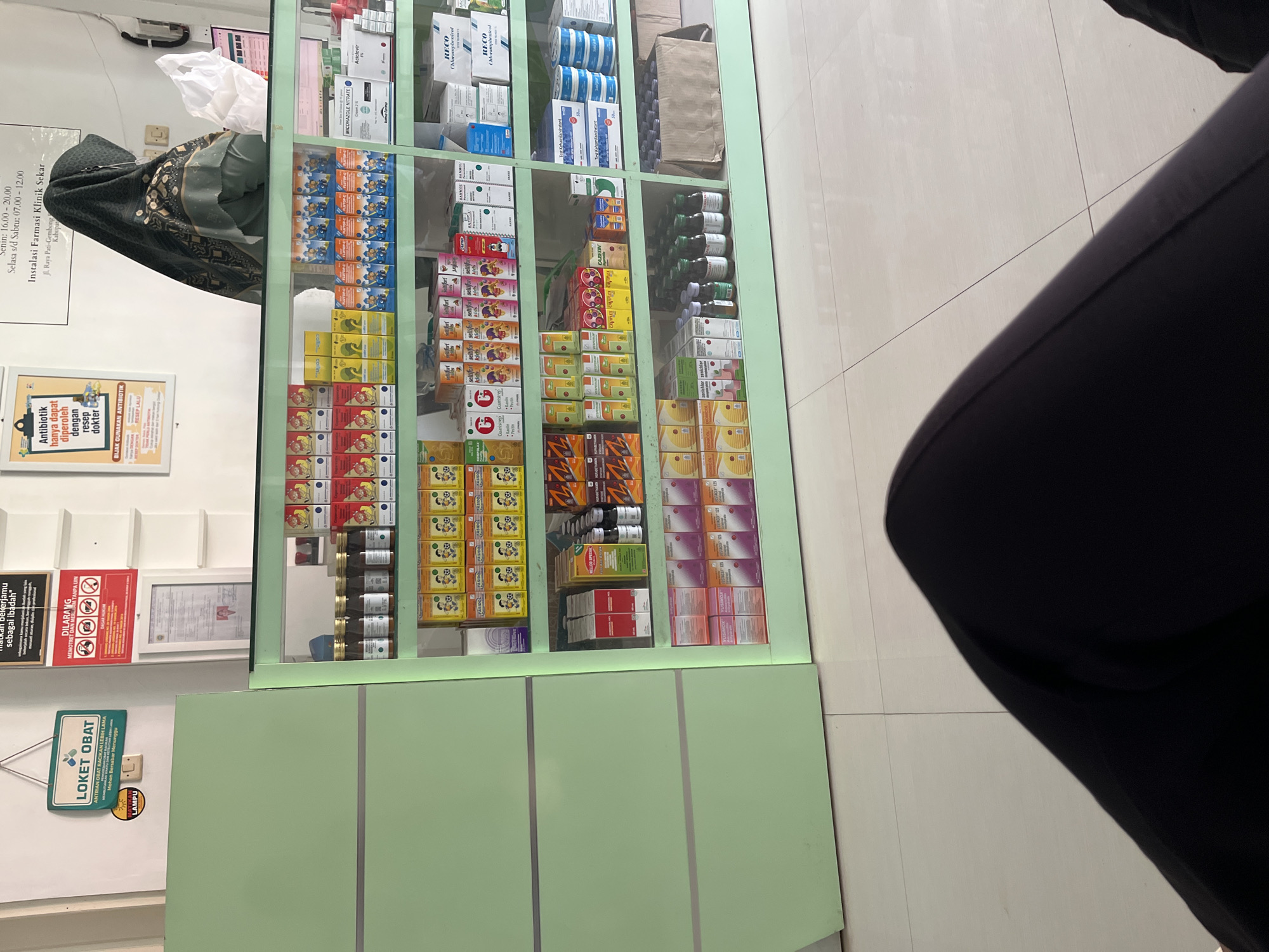 The image shows the interior of a pharmacy in Gembong, Indonesia, with shelves stocked with various medicines. A woman wearing a hijab is visible behind the counter, appearing to be interacting with a customer or organizing inventory. The pharmacy has a clean and organized appearance, with clear signage and displays. The overall impression is that of a functional and well-stocked healthcare facility.