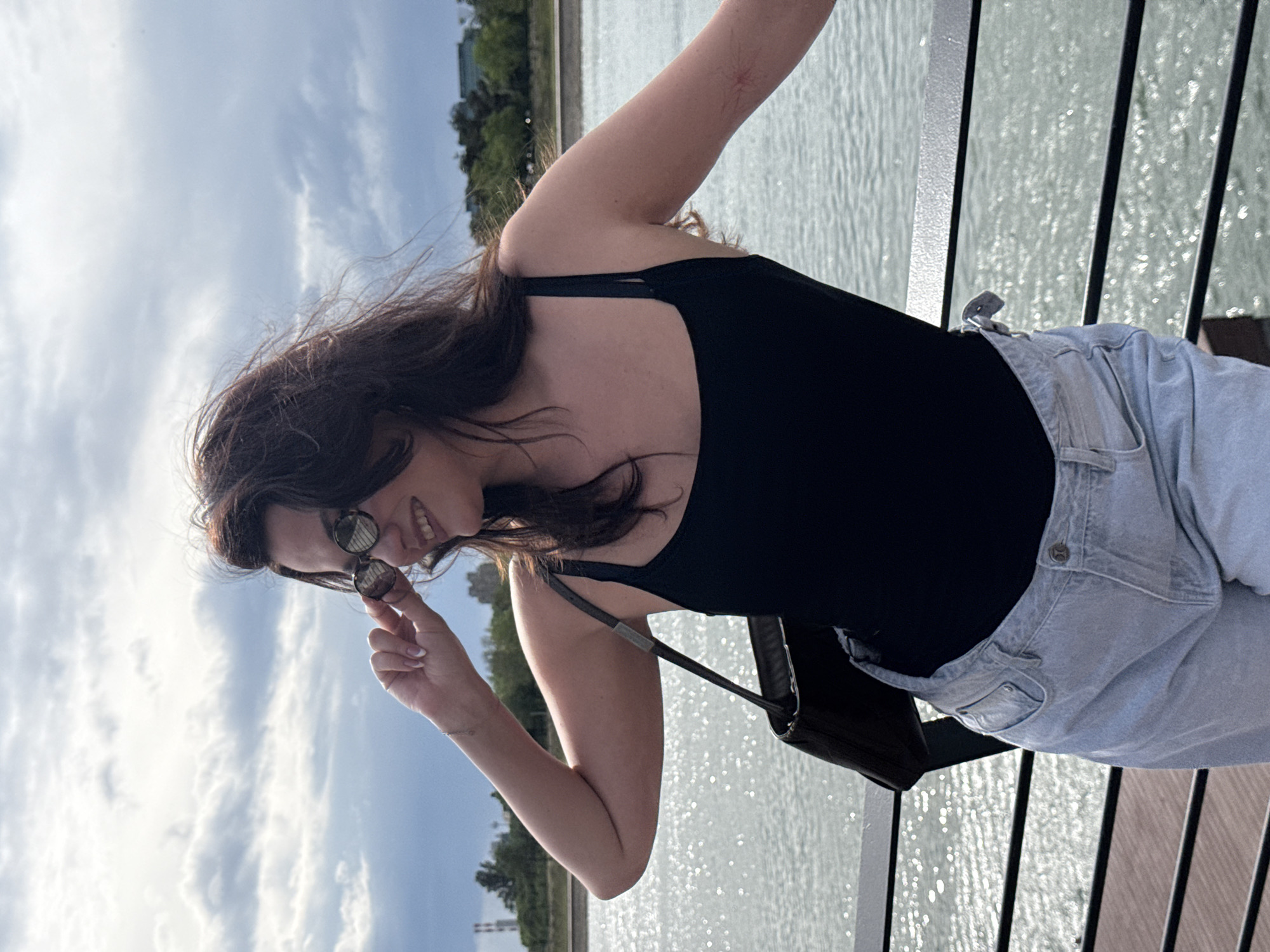 A woman stands on a wooden pier railing, appearing to be leaning over the edge with her right arm resting on the railing. She is dressed in a black tank top and light-colored denim jeans. She is holding a pair of sunglasses in her left hand, partially obscuring her face. A black purse hangs from her shoulder.
The background consists of a body of water and a skyline with buildings and trees. The sky is partly cloudy.
While the exact location cannot be determined with certainty from the image, it is asserted to be in the city of Belgrade, Serbia.