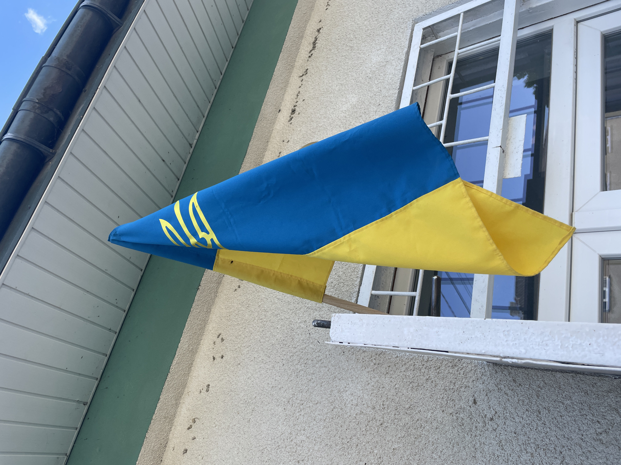 An overhead, upward-angle shot captures a portion of a building's exterior on a clear day, with a flag prominently displayed. The flag, composed of two horizontal stripes, a blue one on top and a yellow one below, is draped over a wooden pole. The blue stripe features a yellow trident emblem. The building's facade is textured and painted in a light, neutral tone, with a contrasting green band running horizontally above it. Above this, a white soffit with visible wooden planks extends outwards, capped by a dark gutter. A white metal railing or frame is visible in the foreground, attached to the wall, with reflections of a blue sky and possibly buildings seen in the window behind it. The scene is located in the city of Shpykolosy, Ukraine.