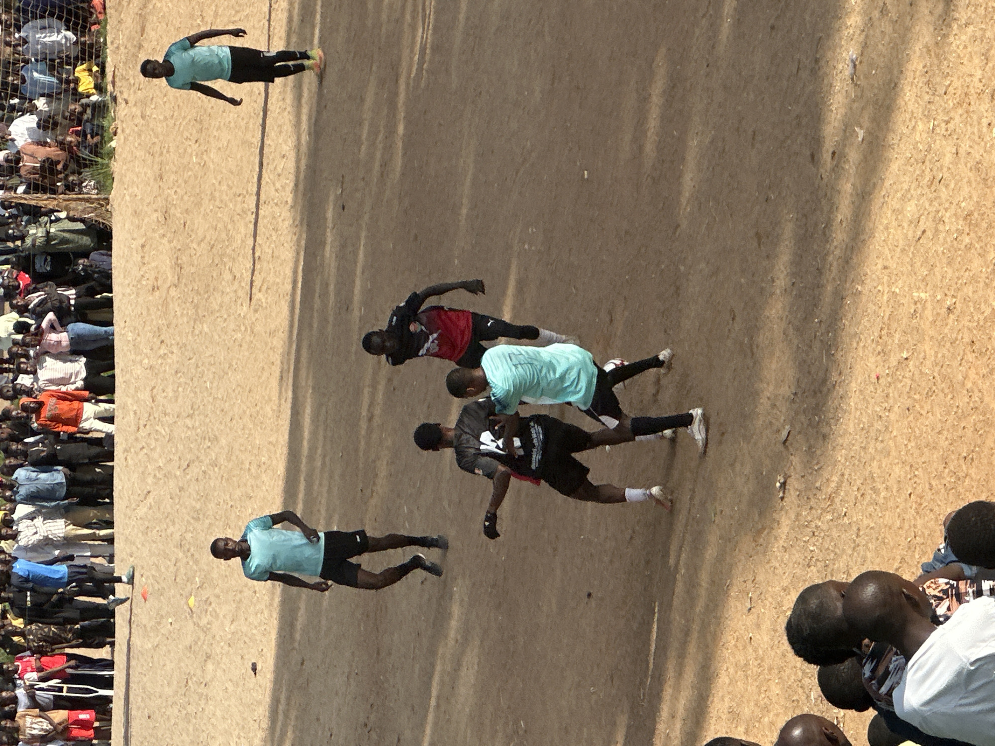 In the city of Jos, Nigeria, a group of young men are engaged in a soccer match on a dirt field. A crowd of onlookers watches from the background. The players are dressed in athletic attire, with some wearing jerseys and shorts, while others are in t-shirts and shorts. The field itself is bare, with no grass, and appears to be uneven. The surrounding area is sandy, with some scattered debris. The overall atmosphere seems to be one of casual competition and community gathering.