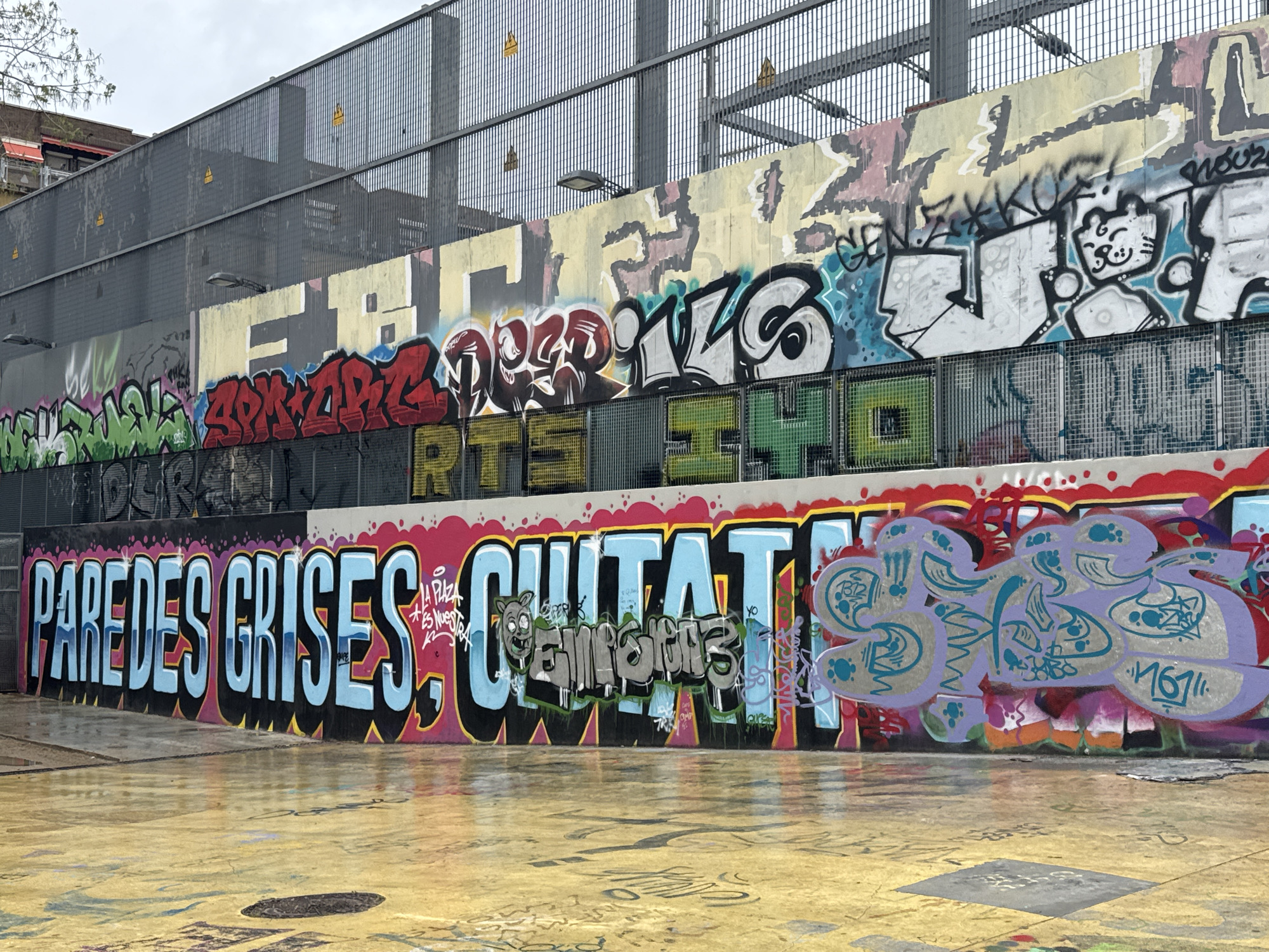 The image shows a graffiti-covered wall in Barcelona, Spain. The graffiti includes stylized lettering, cartoon-like figures, and abstract designs. The wall appears to be part of a multi-level structure with a metal fence above. The ground in front of the wall is wet and also covered in graffiti. The sky is overcast.