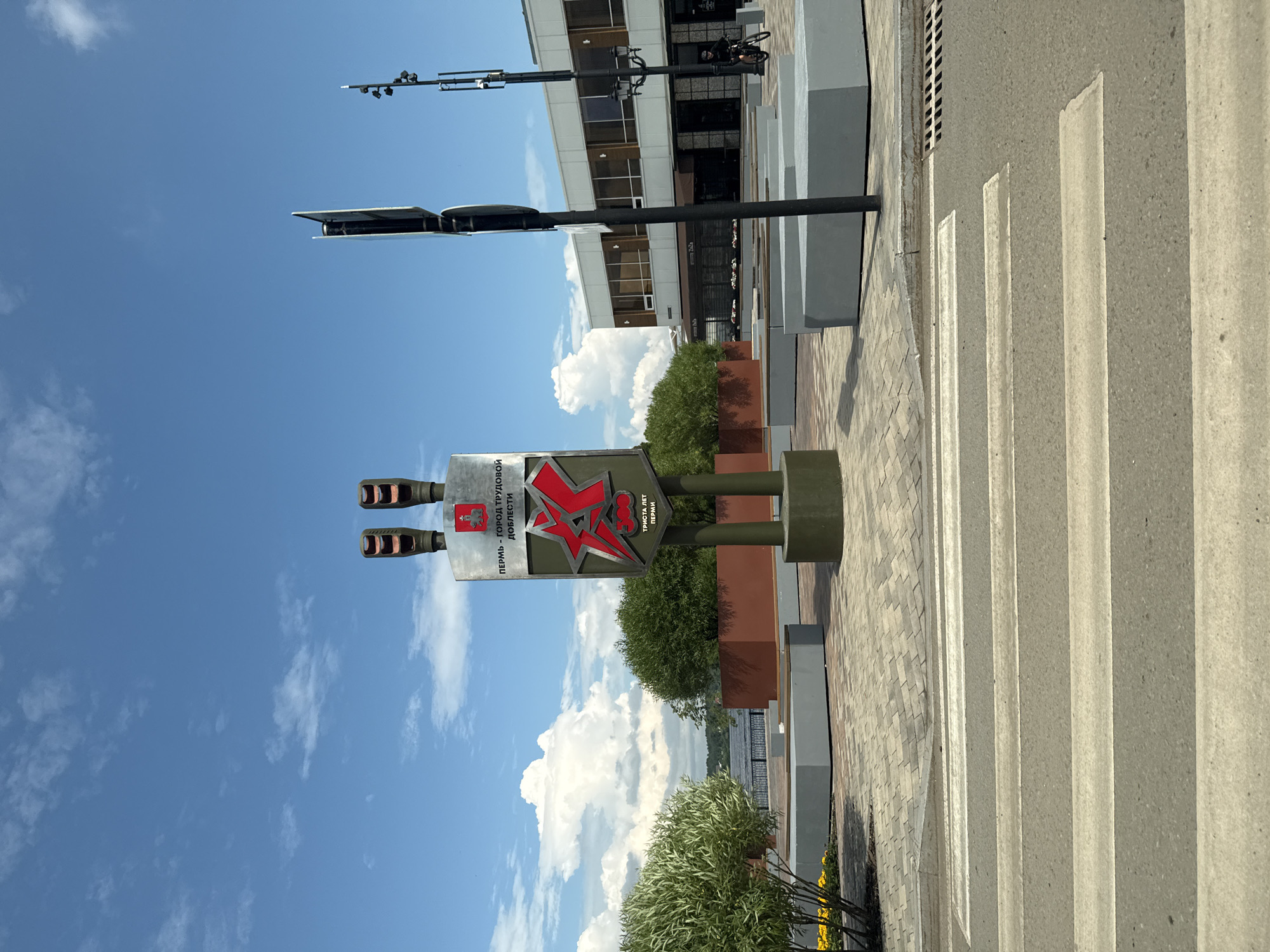 A monument with two traffic light heads on top and a red star emblem on its front, reading "PERM - CITY OF LABOR VALOR" and "300 YEARS OF PERM," stands prominently in a public square in Perm, Russia. The monument is surrounded by modern concrete benches and planters filled with greenery. In the foreground, a pedestrian crossing is visible, and a building with a white facade and dark windows is situated in the background. The sky is blue with scattered clouds, and a body of water can be seen to the left of the monument. A person on a bicycle is riding past the building in the background. The scene is set on a sunny day.