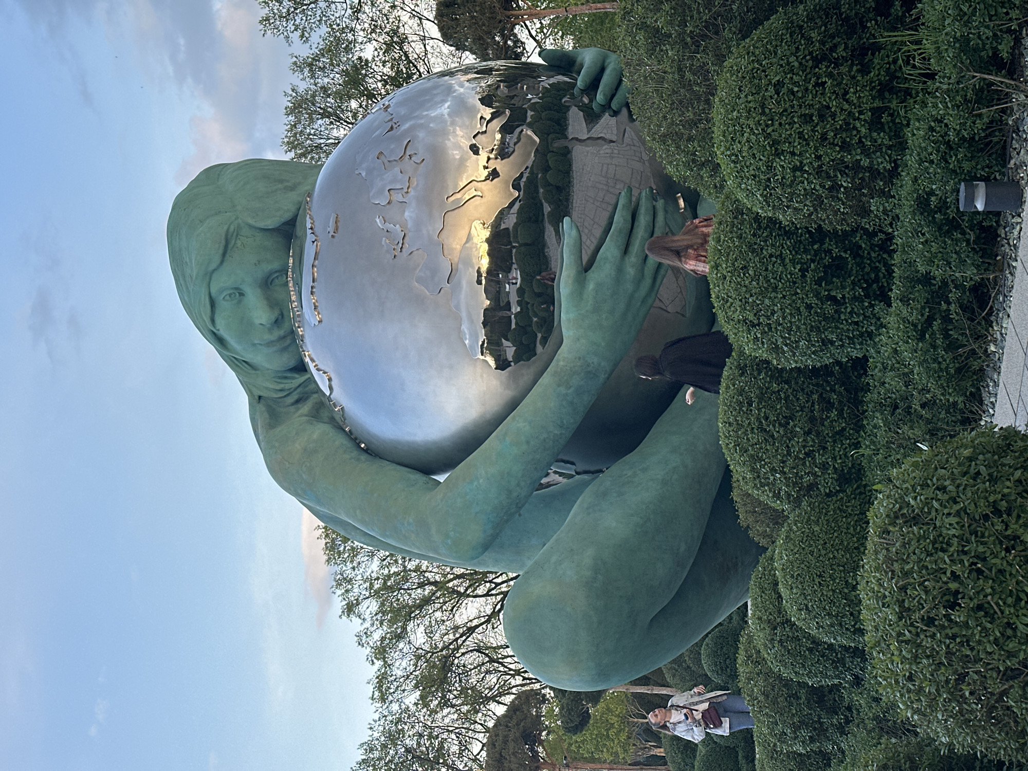 The image shows a statue of a woman in Krasnodar, Russia. The woman is made of a green material and is holding a reflective globe. The continents are cut out of the globe. The statue is surrounded by trees and bushes. There are some people in the background, including a woman with a long hair. The sky is light blue with some clouds.