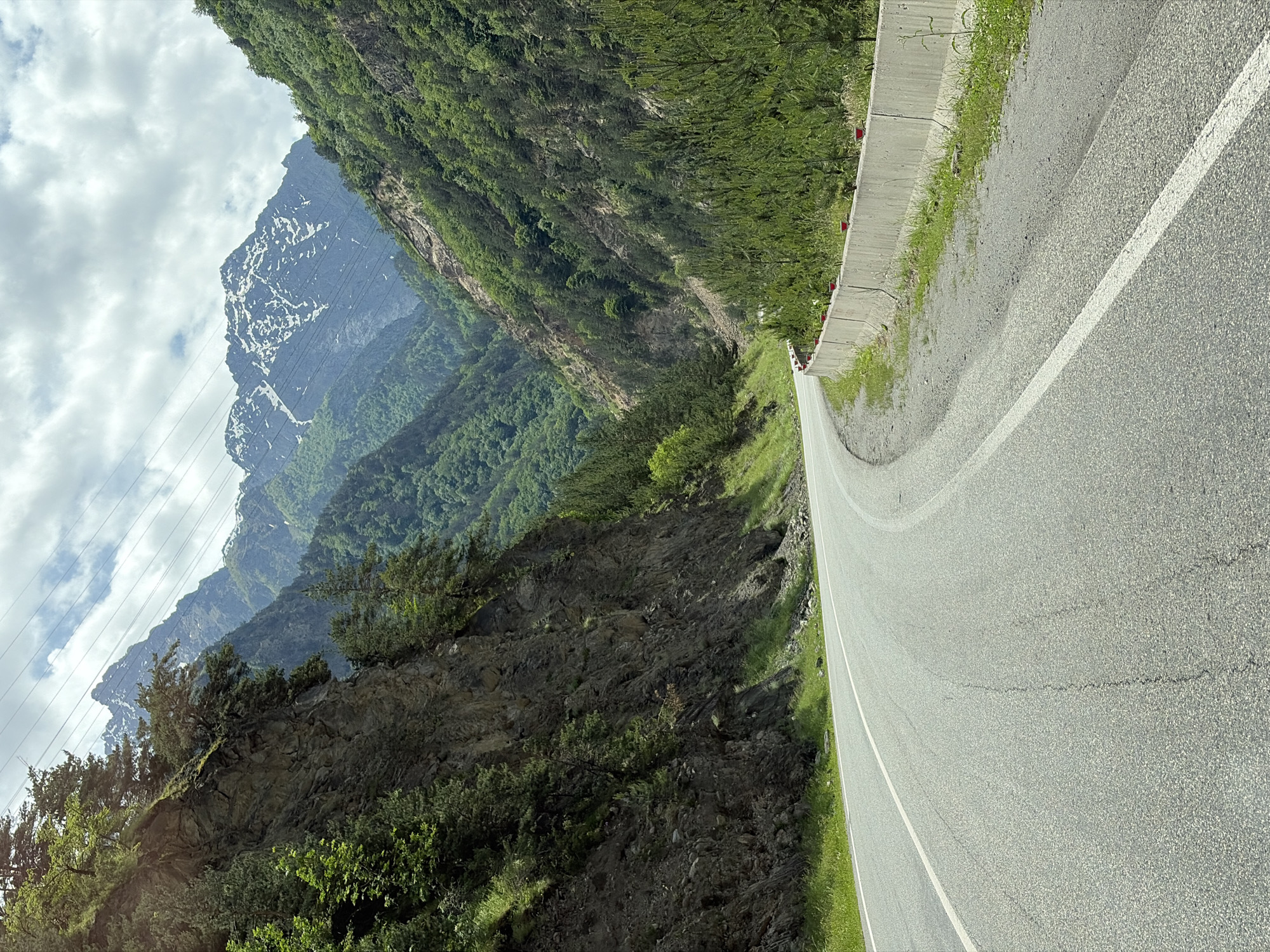 The image depicts a winding asphalt road passing through a mountain valley. The road is bordered by a concrete barrier on the right and a steep, rocky cliff face covered in vegetation on the left. The valley is densely forested with green trees, and in the distance, snow-capped mountains rise against a cloudy sky. Overhead power lines are visible crossing the sky. The scene appears to be located on a mountain pass, possibly in the vicinity of Nuzal, Russia, given the characteristic landscape of the Caucasus Mountains. There are no people or vehicles visible in the image.