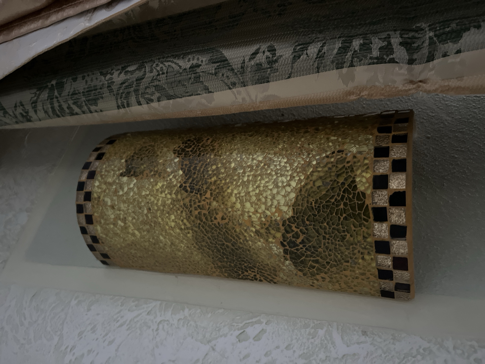 The image shows a cylindrical mosaic lamp with a gold, textured surface, likely made of small, irregular mosaic tiles. The lamp is set into a white niche on a textured white wall. There are black and gold checkered mosaic bands at the top and bottom of the lamp. To the right of the lamp are dark green curtains with a subtle pattern, partially obscuring the wall and the lamp. The lighting is dim, creating shadows. The scene appears to be an interior setting, possibly a hallway or alcove. There are no people or actions depicted. The location in Tarnivtsi, Ukraine, is not evident from the image itself.