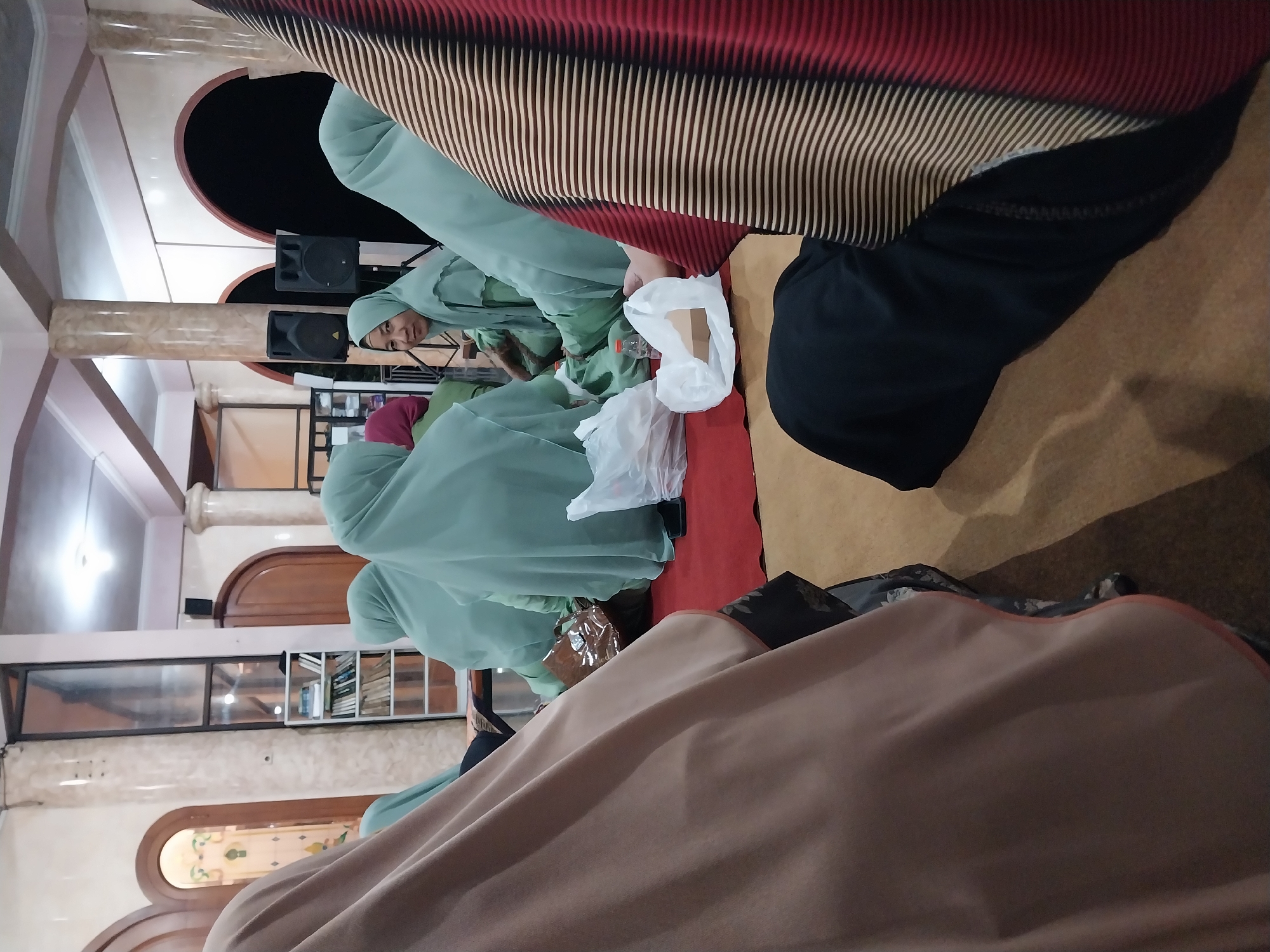 The image shows a group of women wearing light green hijabs, sitting on a red carpet. The women are positioned in what appears to be an indoor space, possibly a mosque or community hall, with a tiled floor visible in the foreground and pillars and arched doorways in the background. There are speakers placed on stands, suggesting a gathering or event. A bookshelf filled with books is visible on the left side of the image. The lighting suggests it is either evening or a dimly lit indoor environment. One woman in the center is looking directly at the camera. The scene is likely located in Nglegok, Indonesia, given the context.