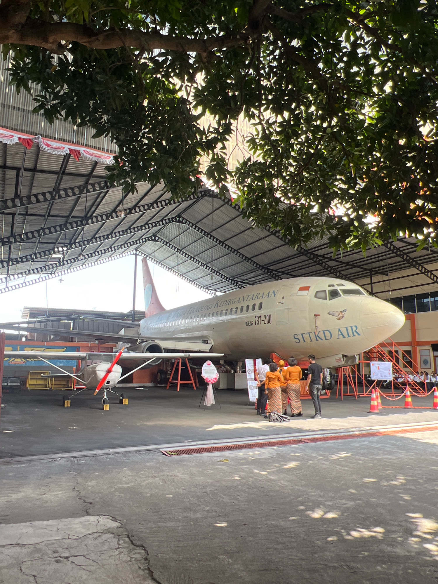 The image depicts an outdoor scene under a covered structure, possibly a hangar or exhibition space, with an airplane as the central subject. The airplane, identified as a Boeing 737-200 with "STTKD AIR" written on its fuselage, is parked on an asphalt surface. Several people, some in traditional Indonesian attire, are standing near the front of the airplane. A smaller aircraft with a red propeller is also visible to the left of the main airplane. The environment appears to be a public area, perhaps a museum or an event venue, with flags and safety cones present. The location is identified as Sewon, Indonesia.