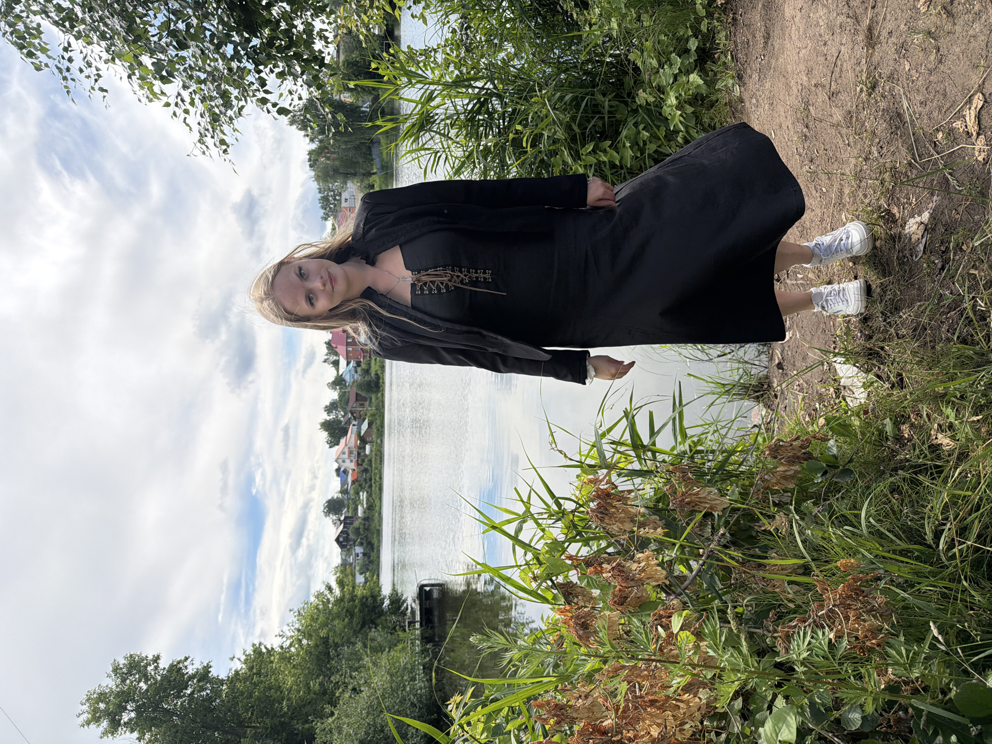 A young woman with blonde hair stands on a dirt path next to a body of water, facing the camera. She is wearing a long black dress with a lace-up front and a black jacket over it. She has on white sneakers. The background shows a calm lake, lush green vegetation along the shore, and a small village with colorful houses in the distance. The sky is partly cloudy. The scene appears to be a serene natural setting on the outskirts of a residential area, possibly in the city of Chaikovsky, Russia, given the architecture and landscape.